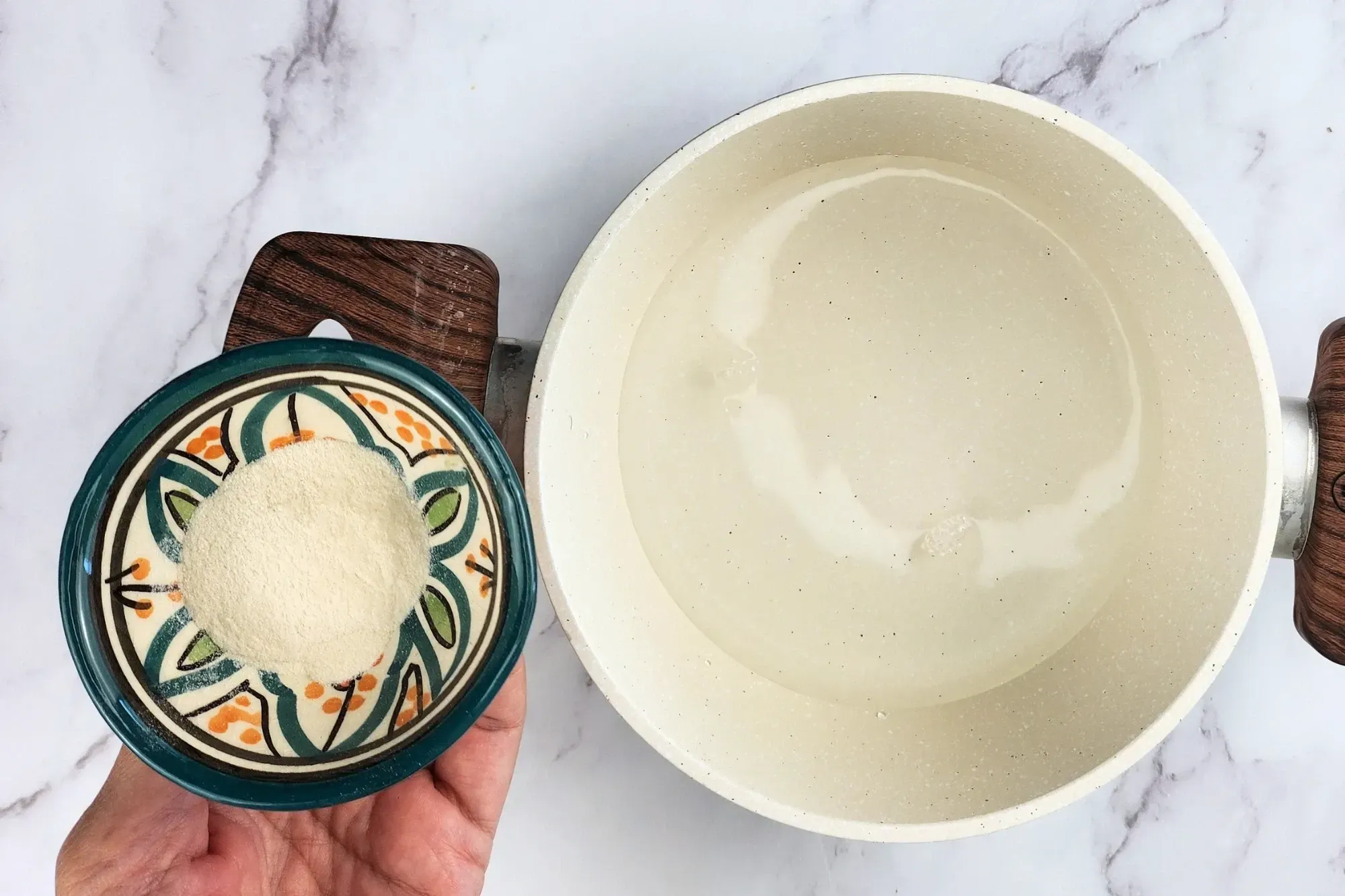 A hand holding a bowl of agar powder next to a pot of water