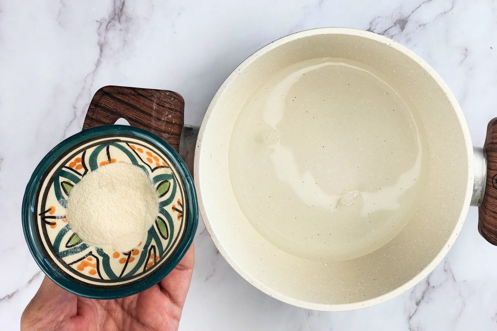 A hand holding a bowl of agar powder next to a pot of water