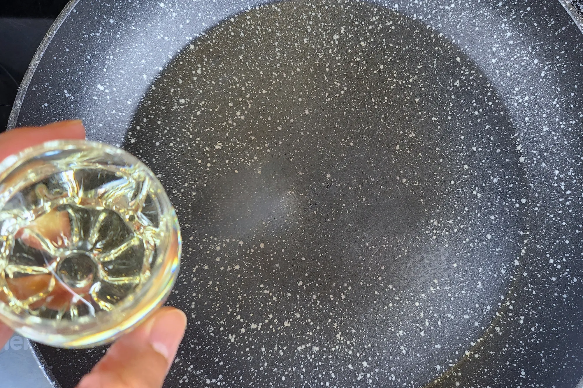 A hand holding a bowl of oil above a frying pan