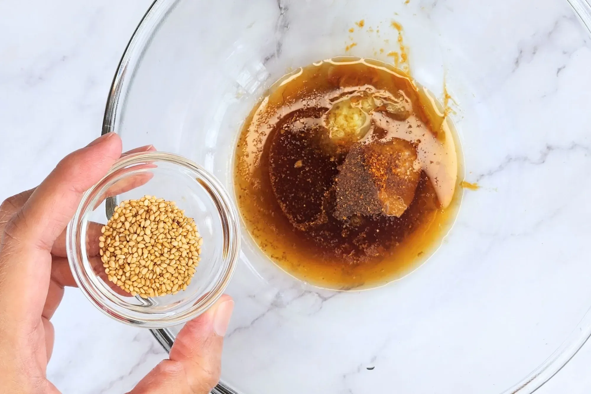A hand holding a bowl of sesame seeds above the dressing bowl