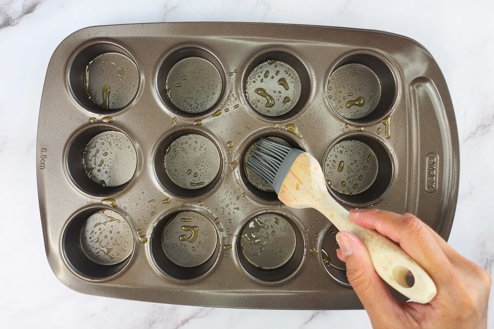 A hand is brushing oil on a muffin tray. 
