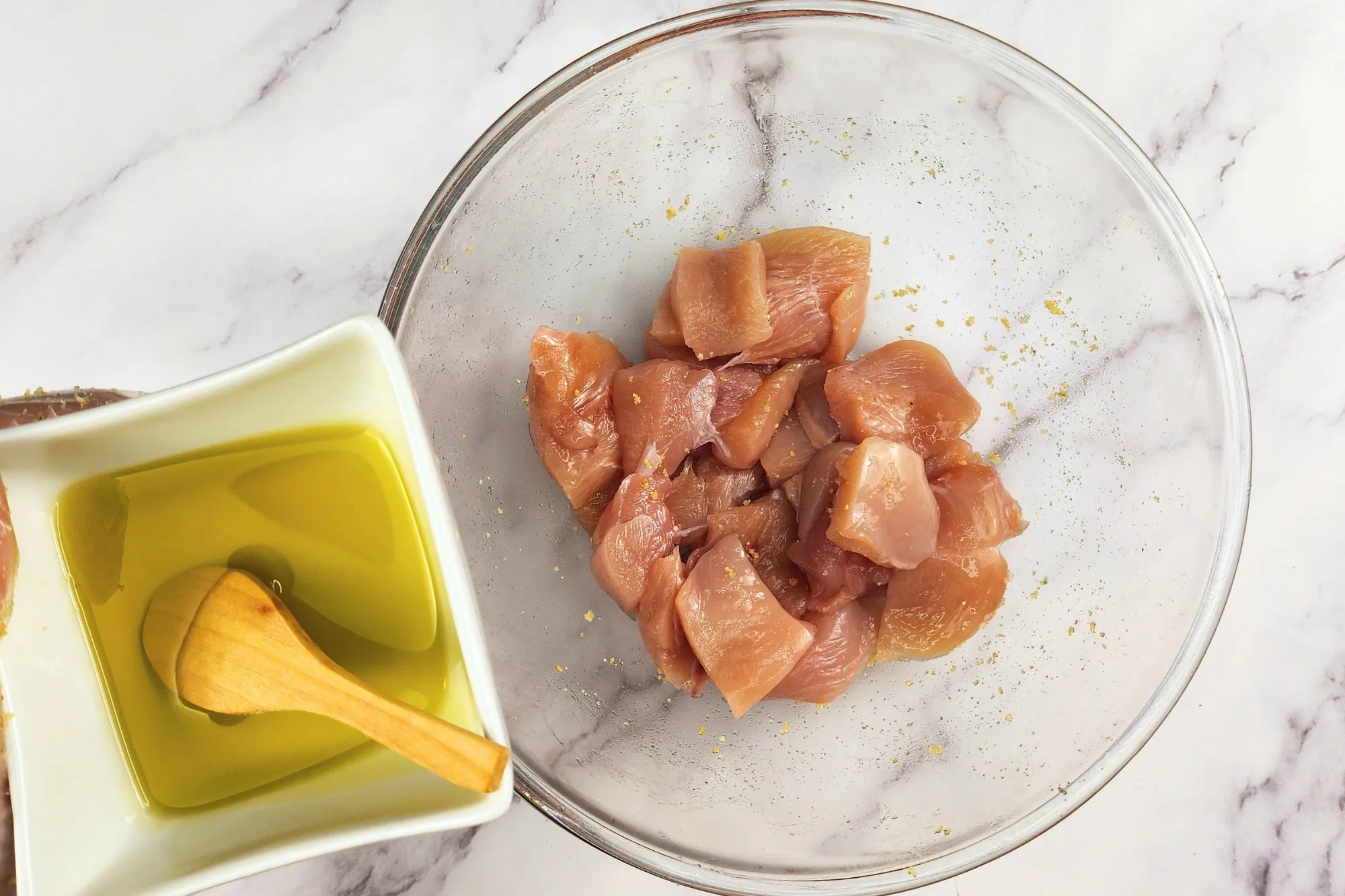 A hand is hold a bowl of olive oil above a mixing bowl with chicken..