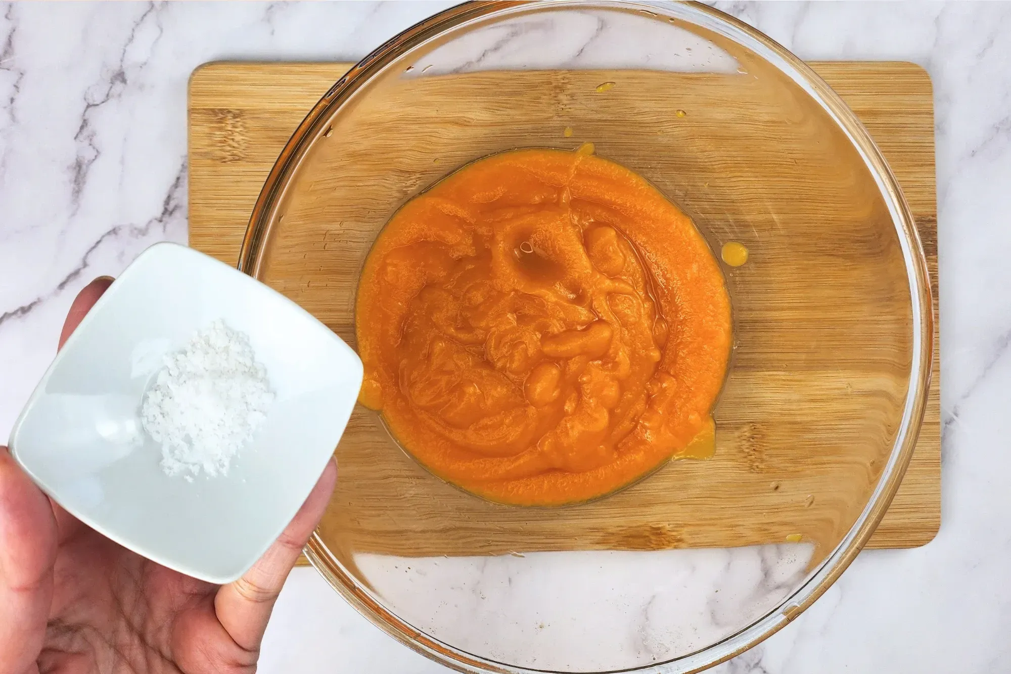 A hand is hold a bowl of salt above a mixing bowl of pumpkin puree