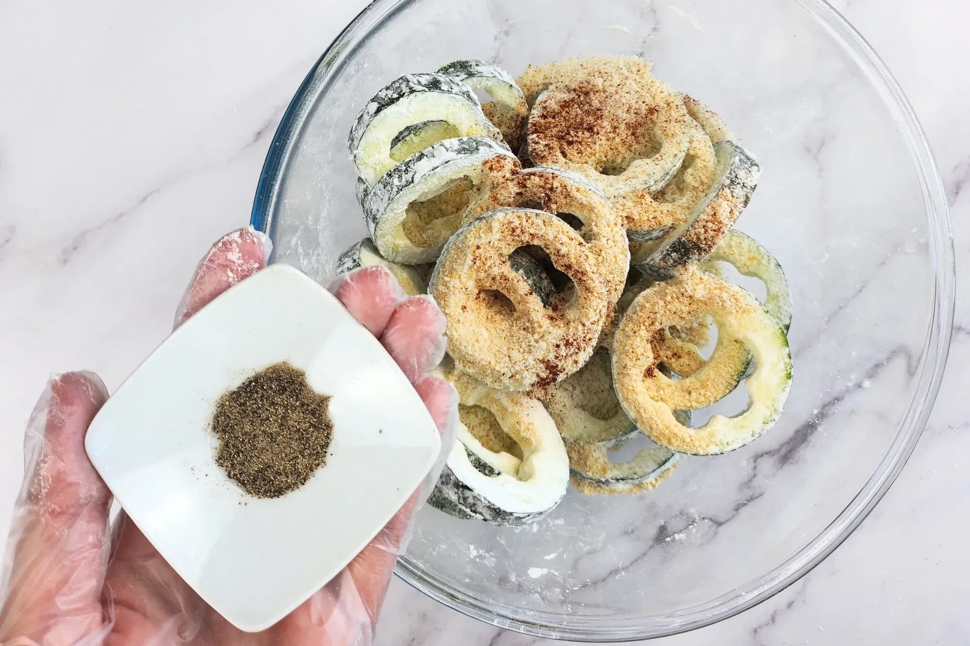 A hand is holding a bowl of black pepper above a bowl of zucchini rings