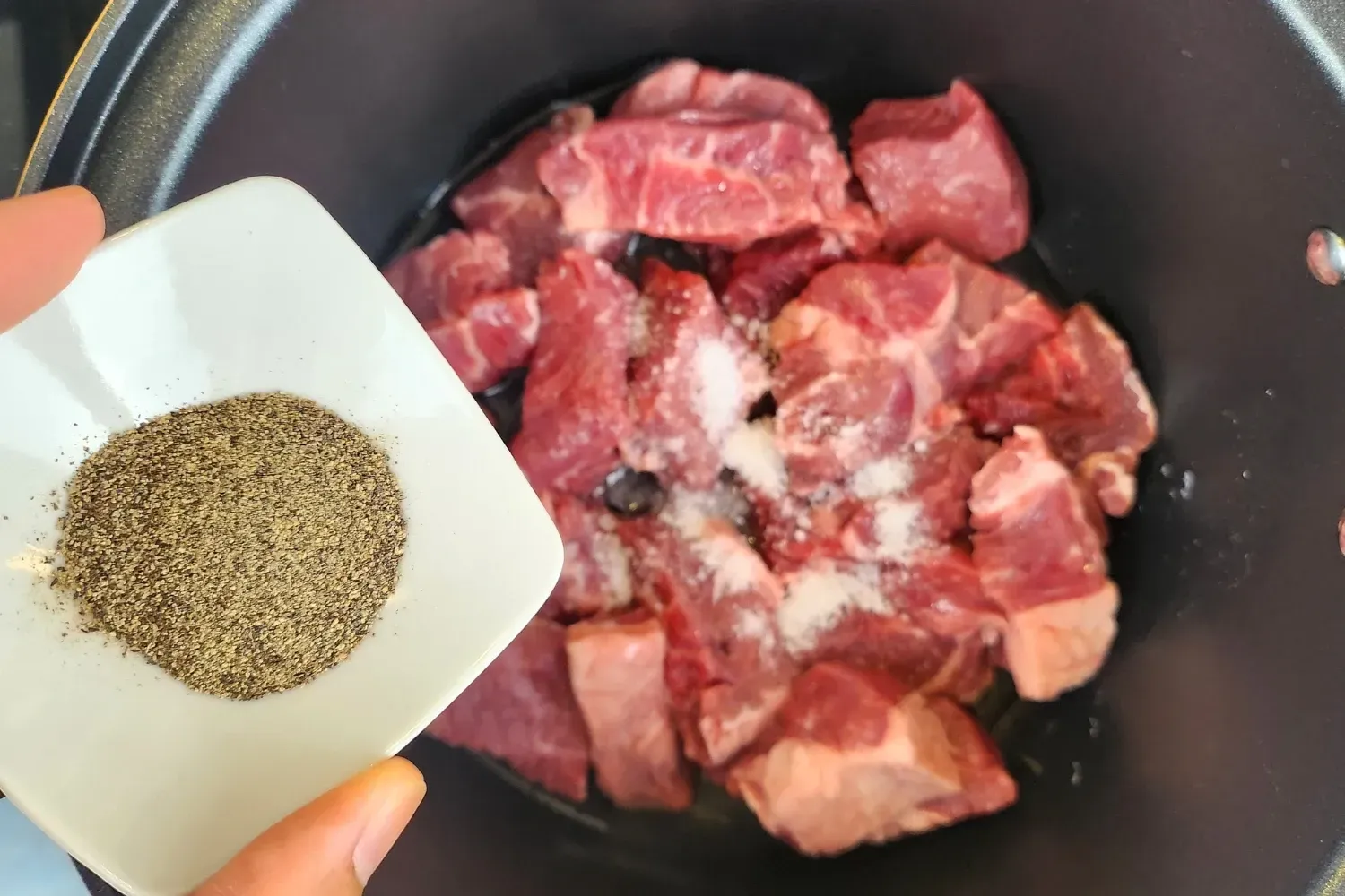 A hand is holding a bowl of black pepper above a pot with meat.