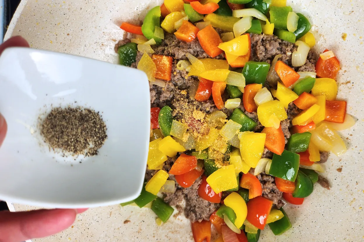 A hand is holding a bowl of black pepper powder above cooked food in a wok.