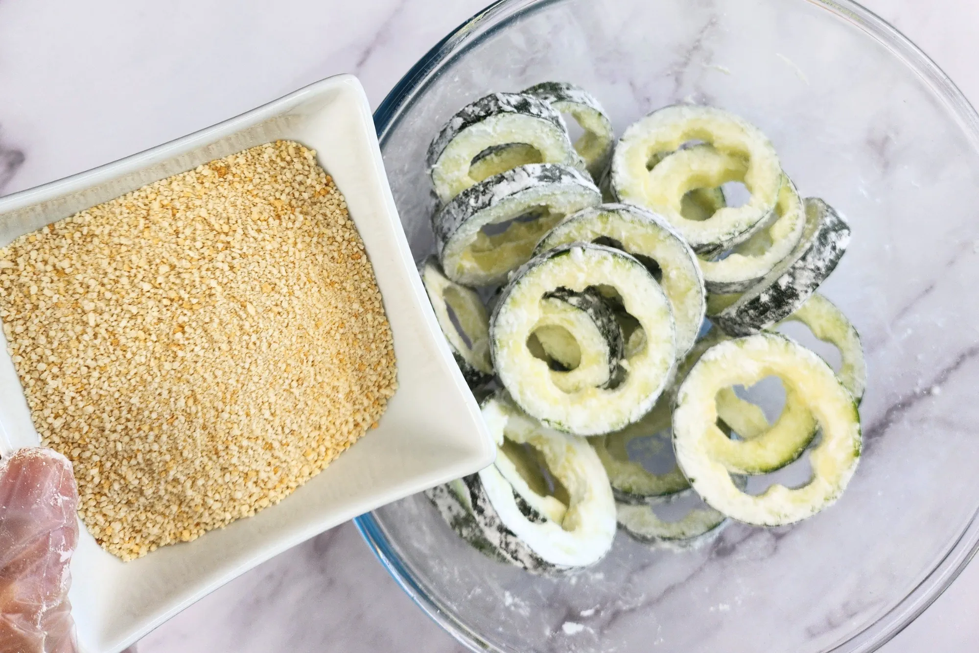 A hand is holding a bowl of breadcrumbs above a bowl of zucchini rings