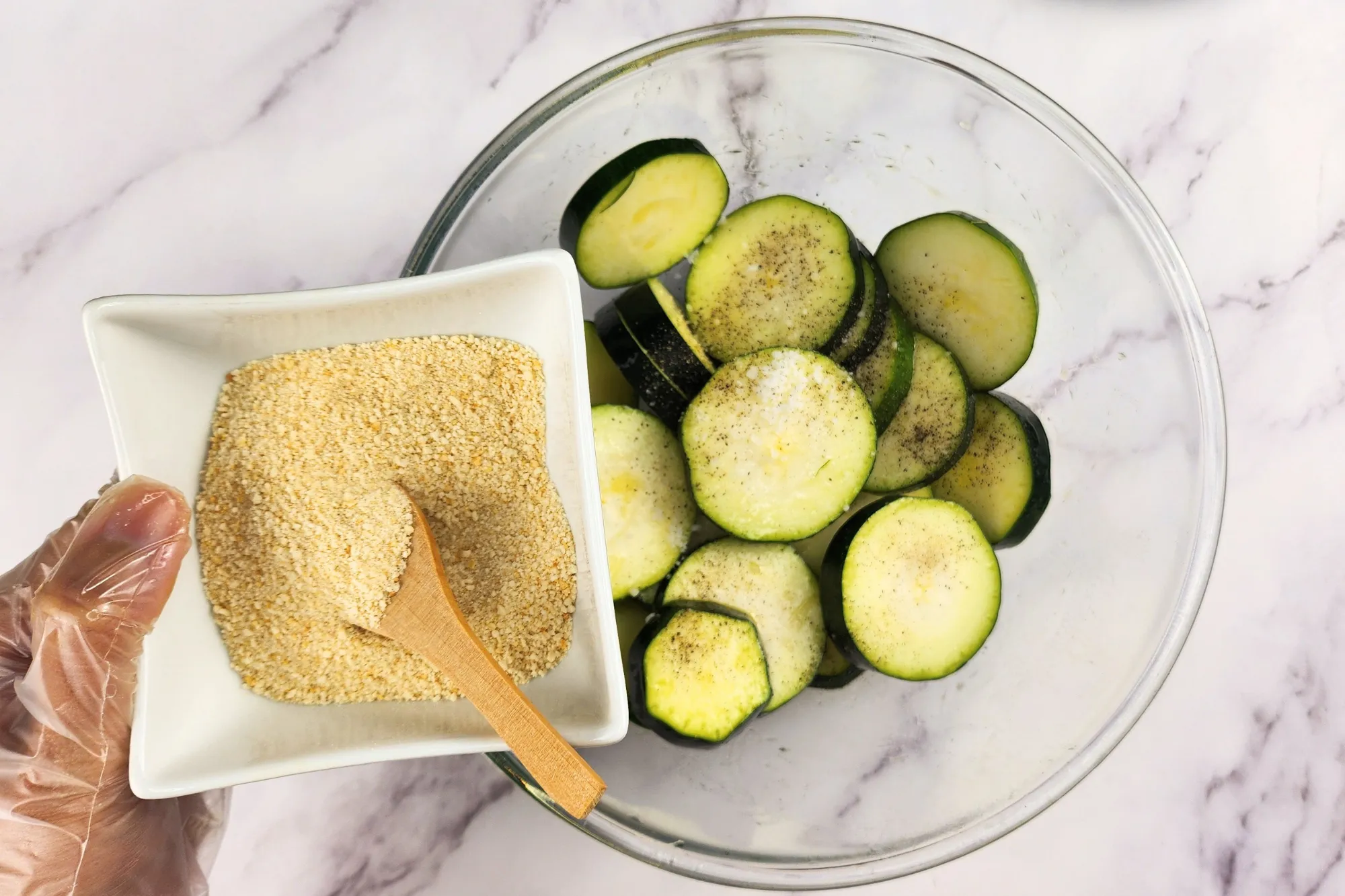 A hand is holding a bowl of breadcrumbs above a mixing bowl with zucchinis. 