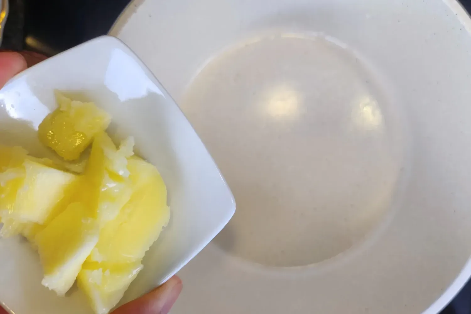 A hand is holding a bowl of butter above a pot.