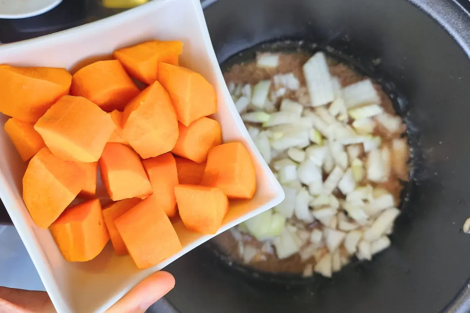 A hand is holding a bowl of carrots above a pot with cooking onions.