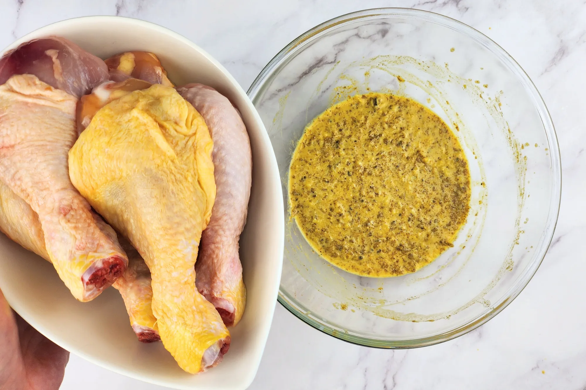 A hand is holding a bowl of chicken drumsticks above a mixing bowl with yellow sauce.