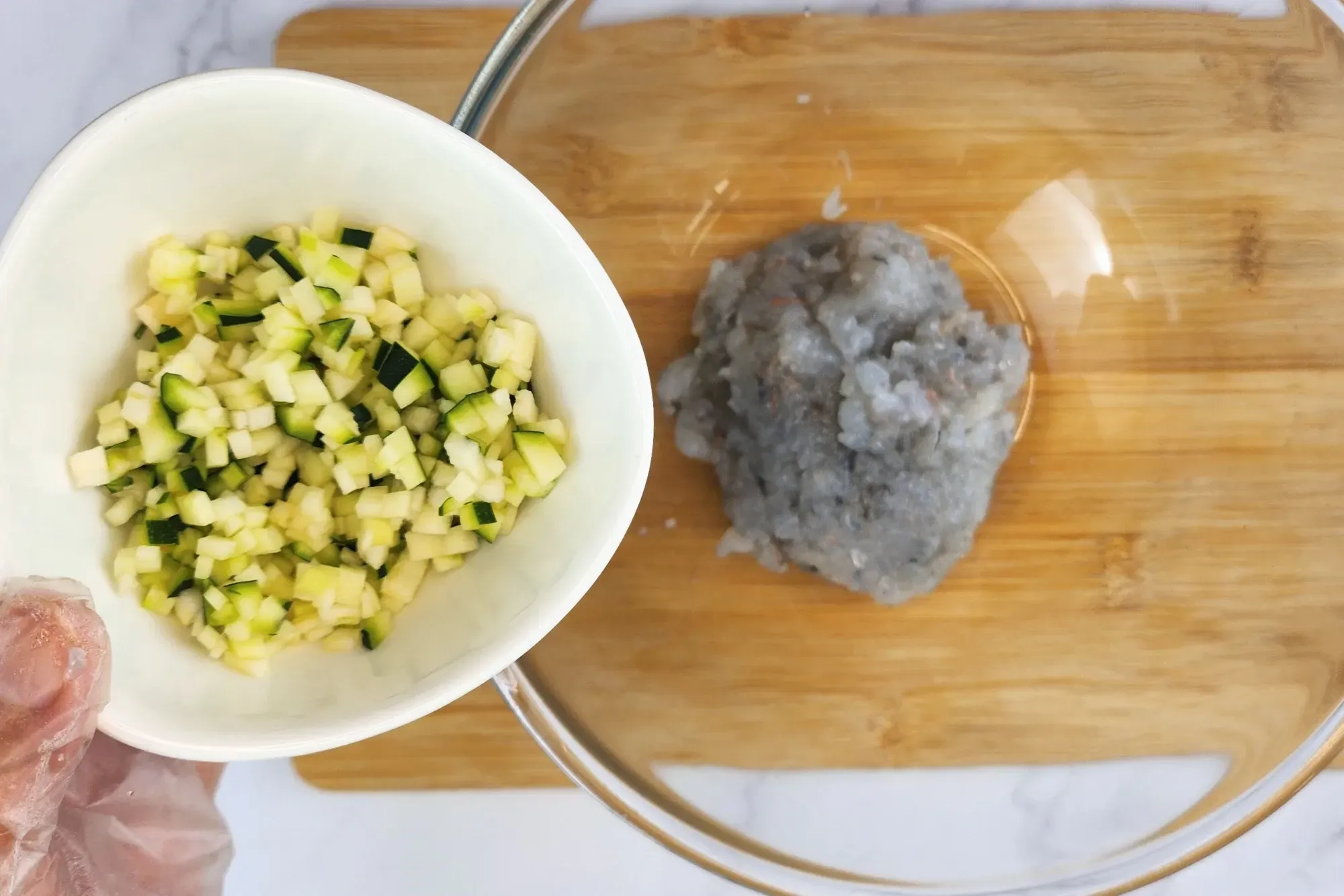 A hand is holding a bowl of chopped zuchini above a mixing bowl