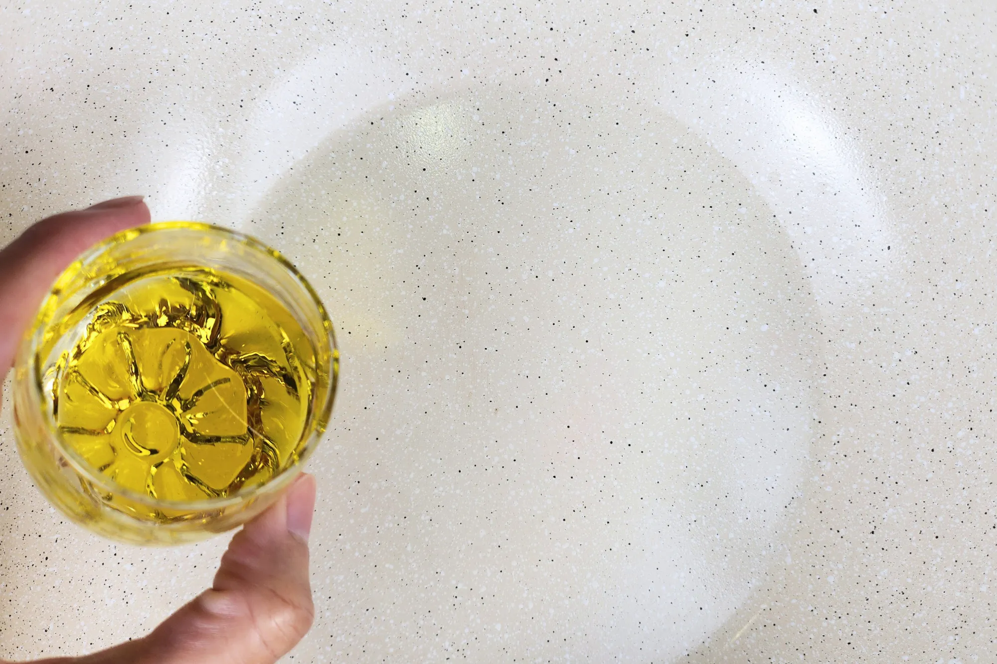 A hand is holding a bowl of cooking oil above a hot wok.