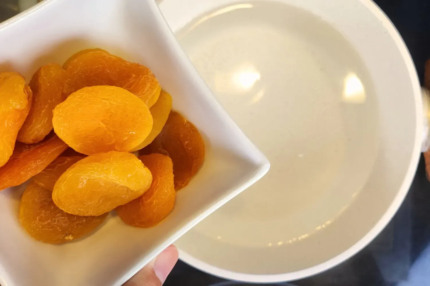 A hand is holding a bowl of dried apricots above a boiling water pot..