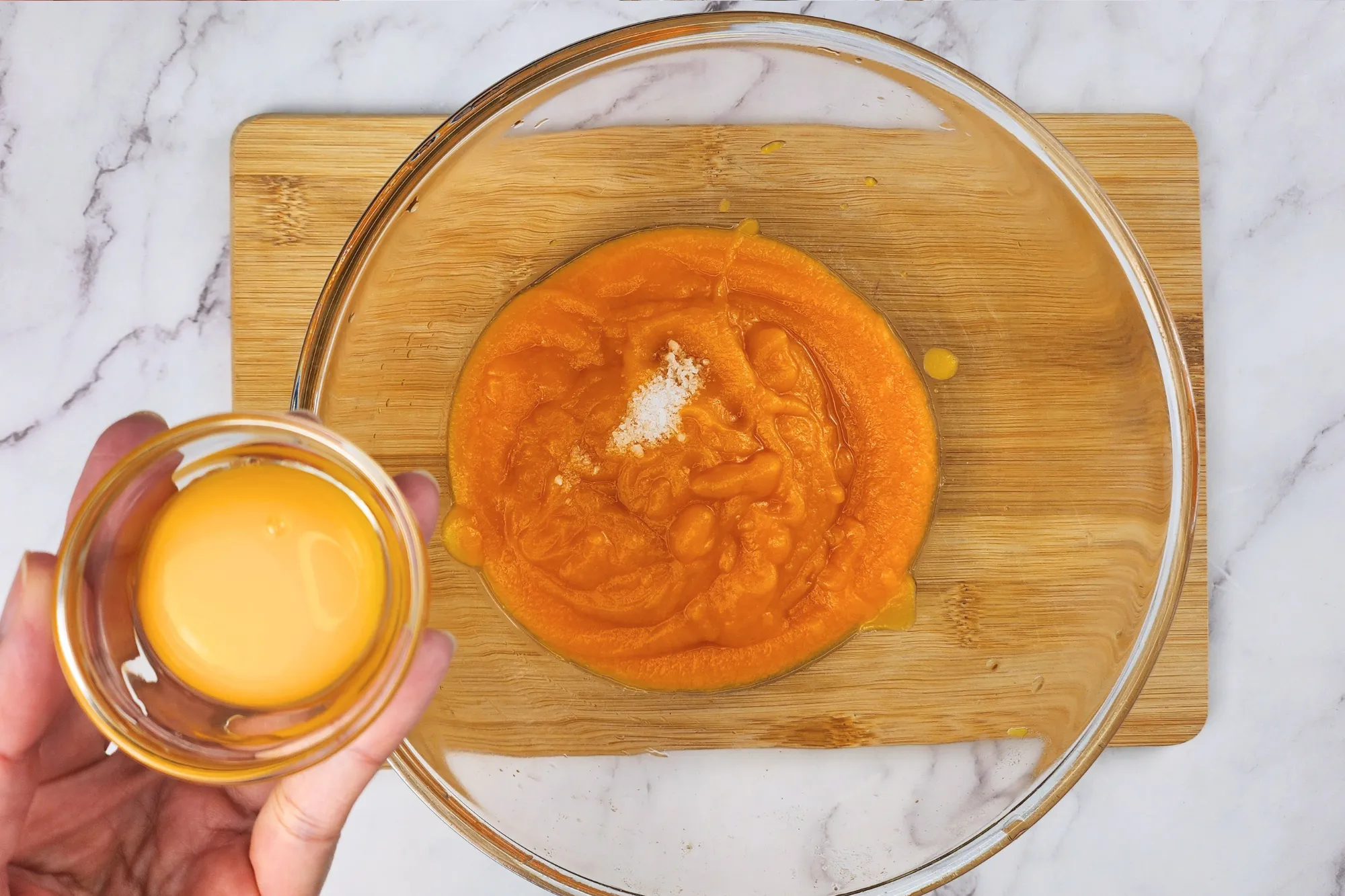 A hand is holding a bowl of egg yolk above a mixing bowl