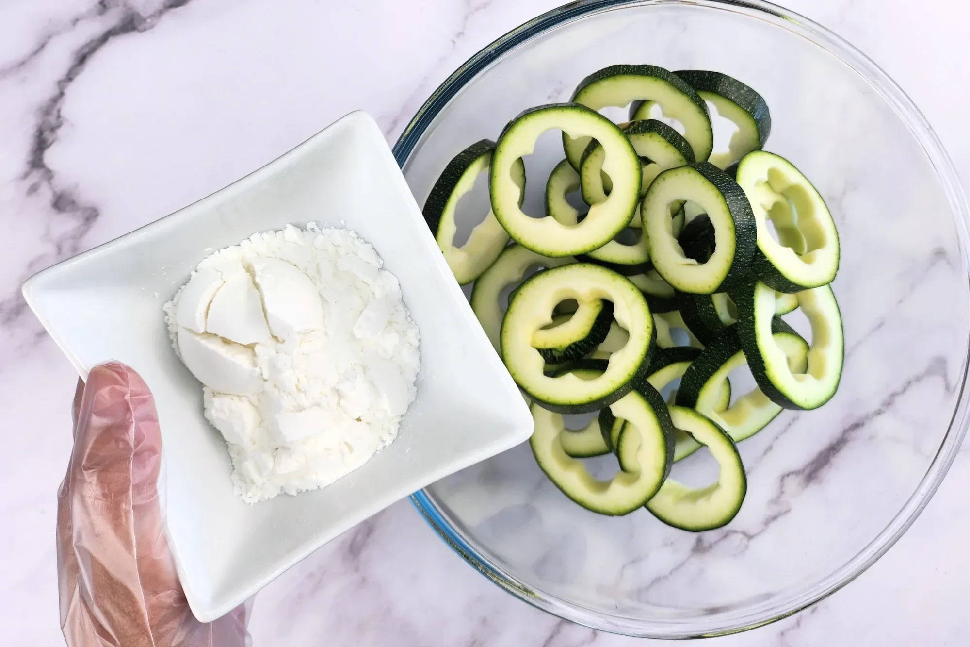 A hand is holding a bowl of flour above a bowl of zucchini rings