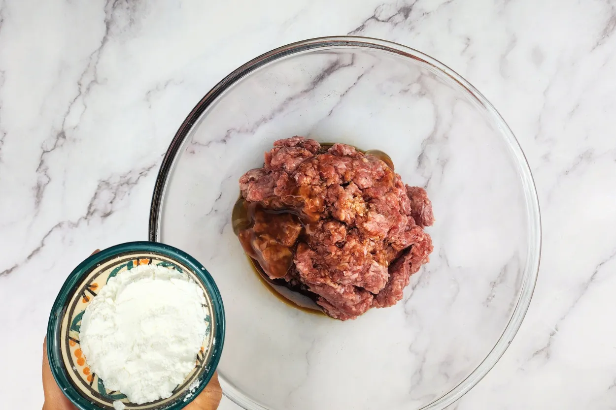 A hand is holding a bowl of flour above a mixing bowl with ground beef.