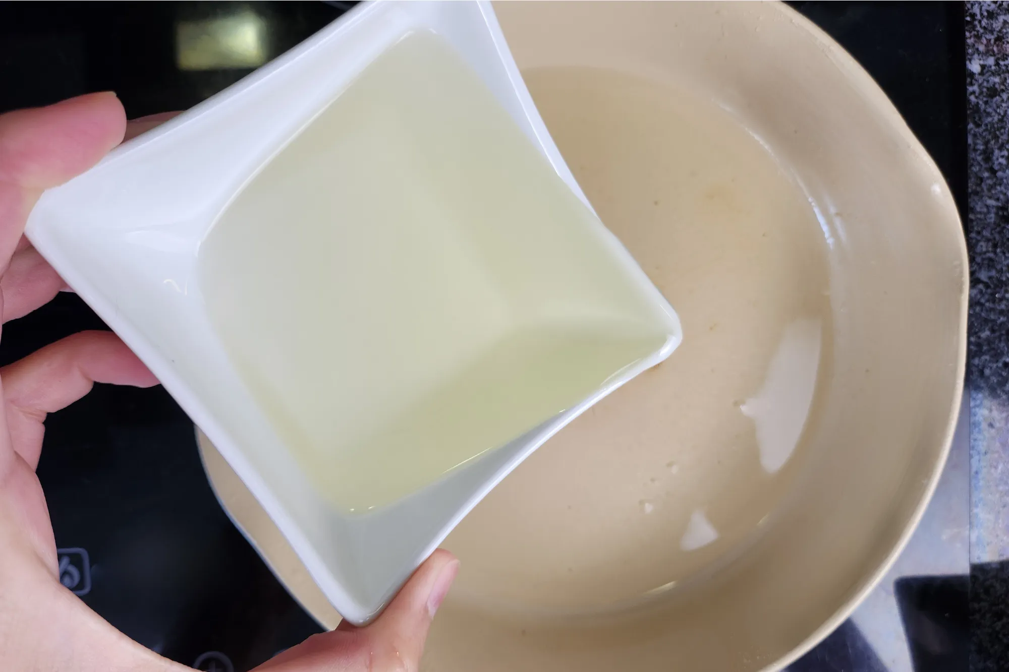 A hand is holding a bowl of frying oil above a sauce pan.