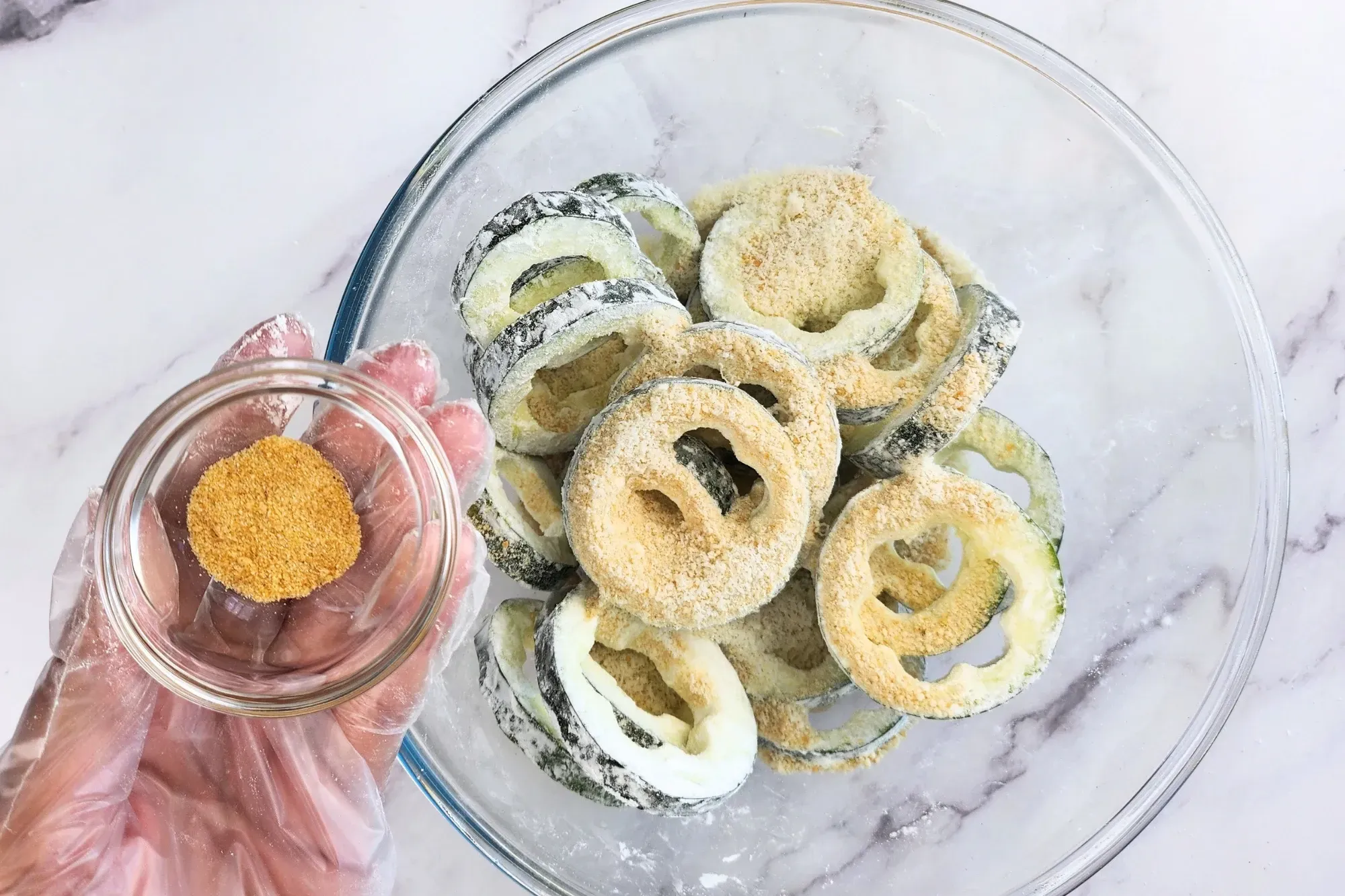 A hand is holding a bowl of garlic powder above a bowl of zucchini rings