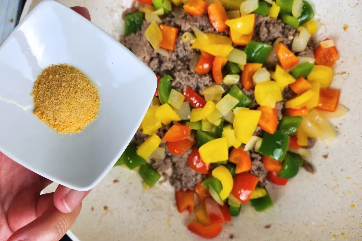A hand is holding a bowl of garlic powder above cooked food in a wok.
