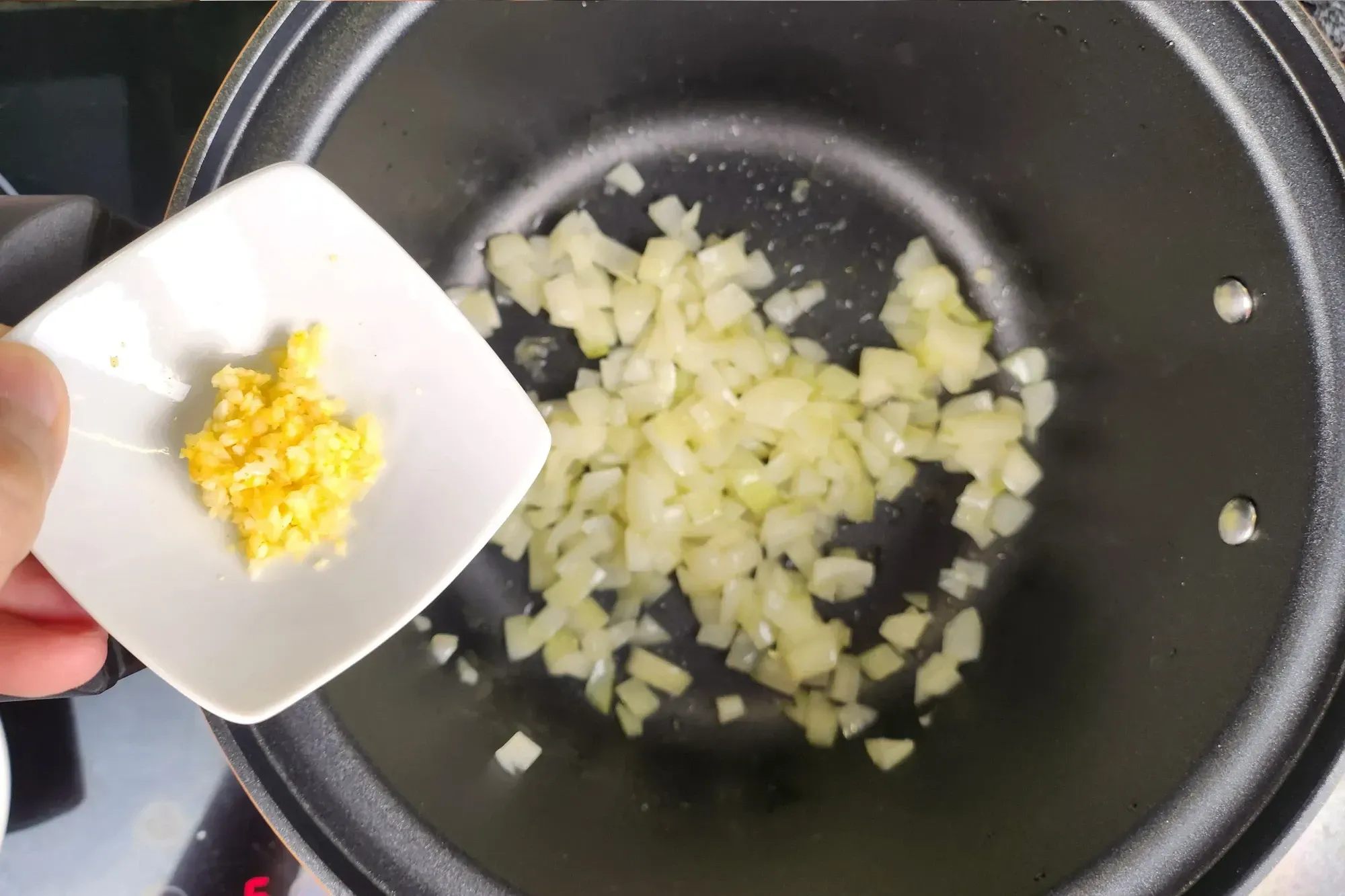 A hand is holding a bowl of grated ginger above a black pot with cooked onion