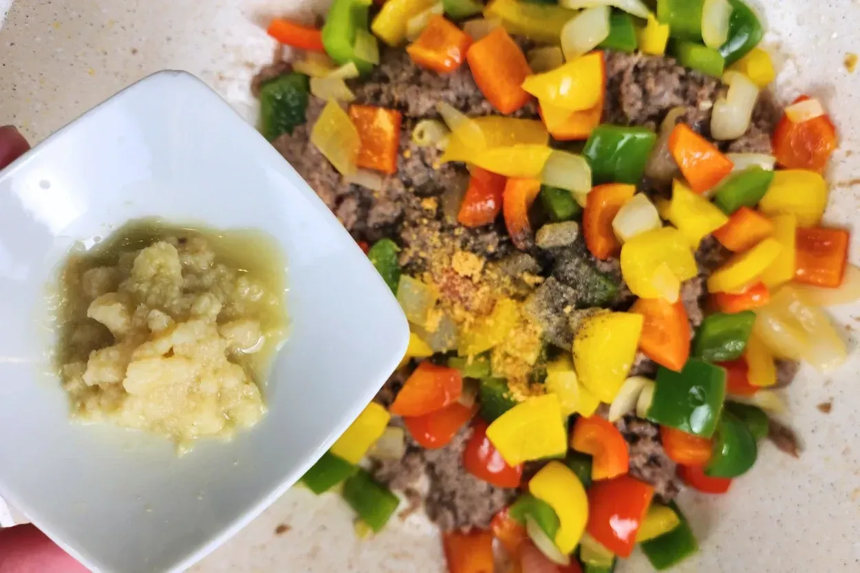 A hand is holding a bowl of grated ginger above cooked food in a wok.