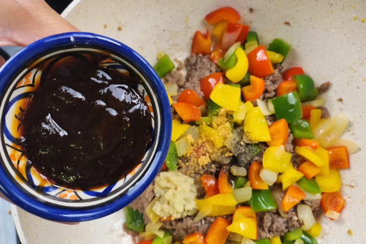 A hand is holding a bowl of hoisin sauce above cooked food in a wok.
