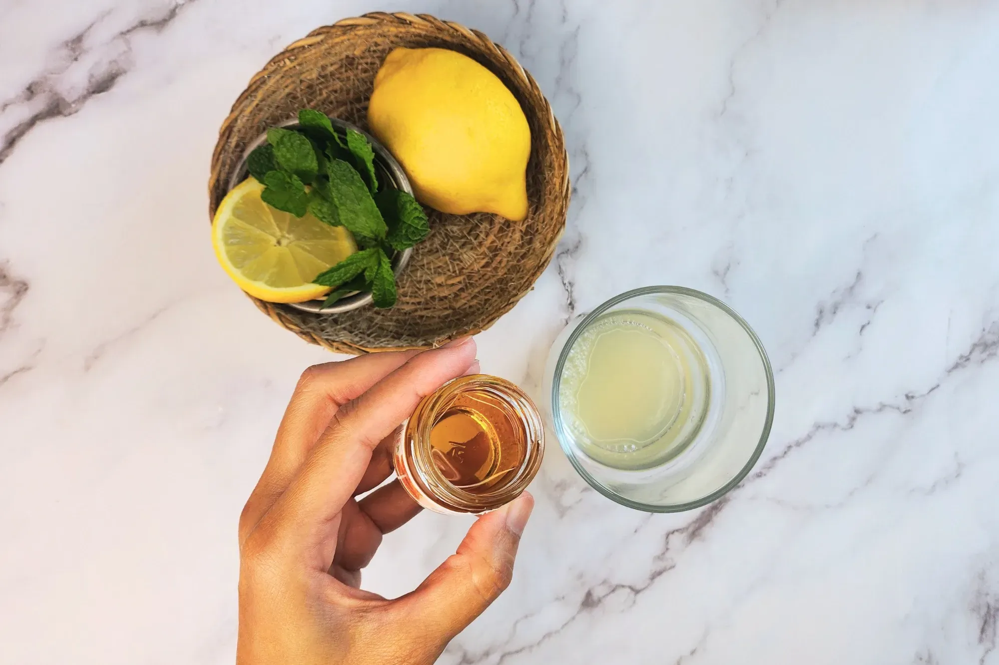 A hand is holding a bowl of honey above a glass next to lemon and mint.