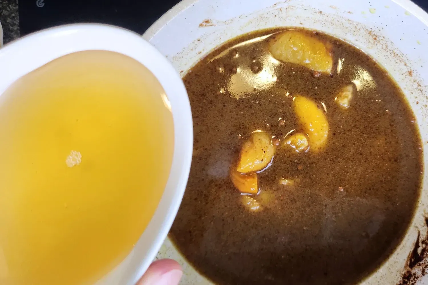 A hand is holding a bowl of honey above a pot with broth and apricots..