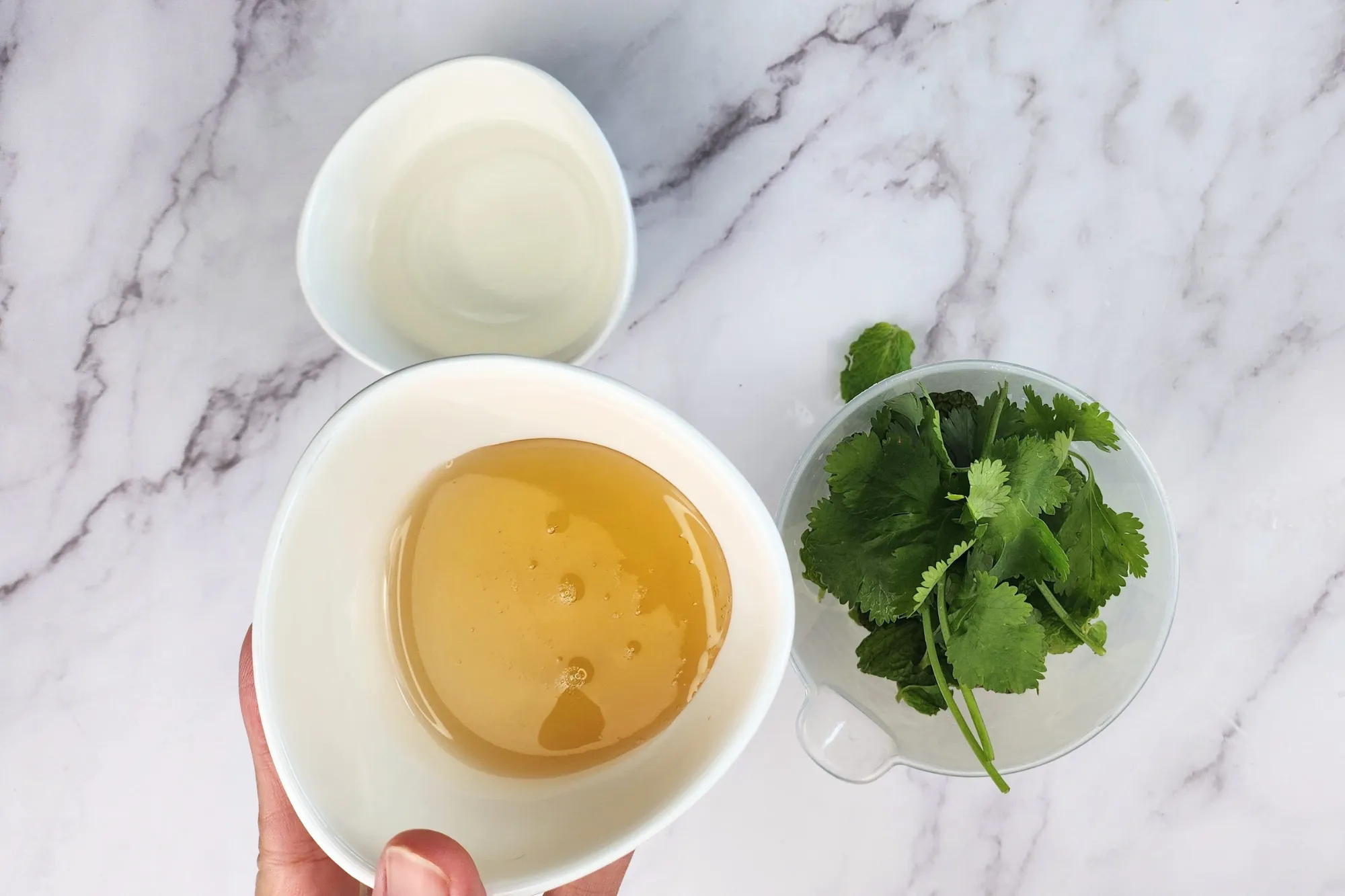 A hand is holding a bowl of honey is above a mixing jar next to a bowl of vinegar.