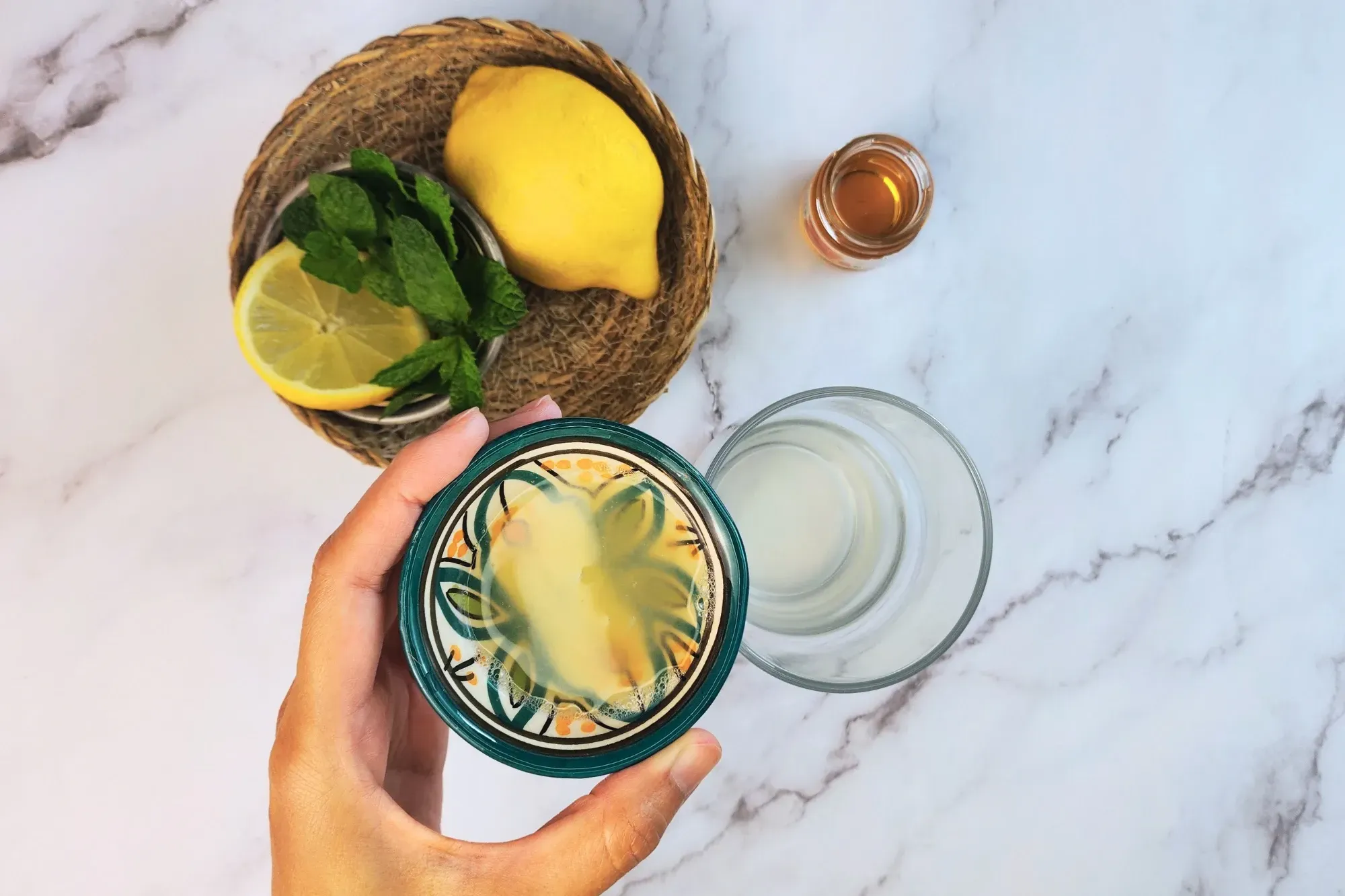 A hand is holding a bowl of lemon juice above a glass next to lemon and mint.