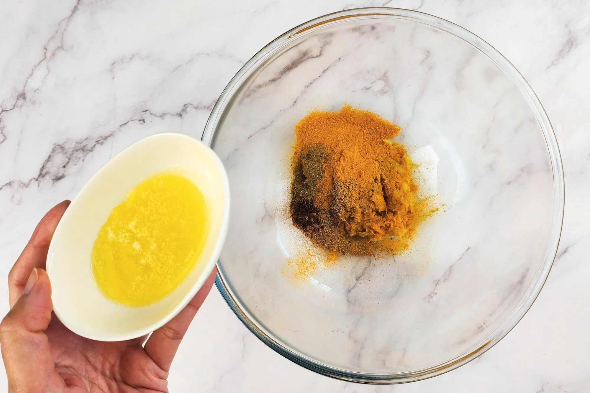 A hand is holding a bowl of melted butter above a mixing bowl with spices.