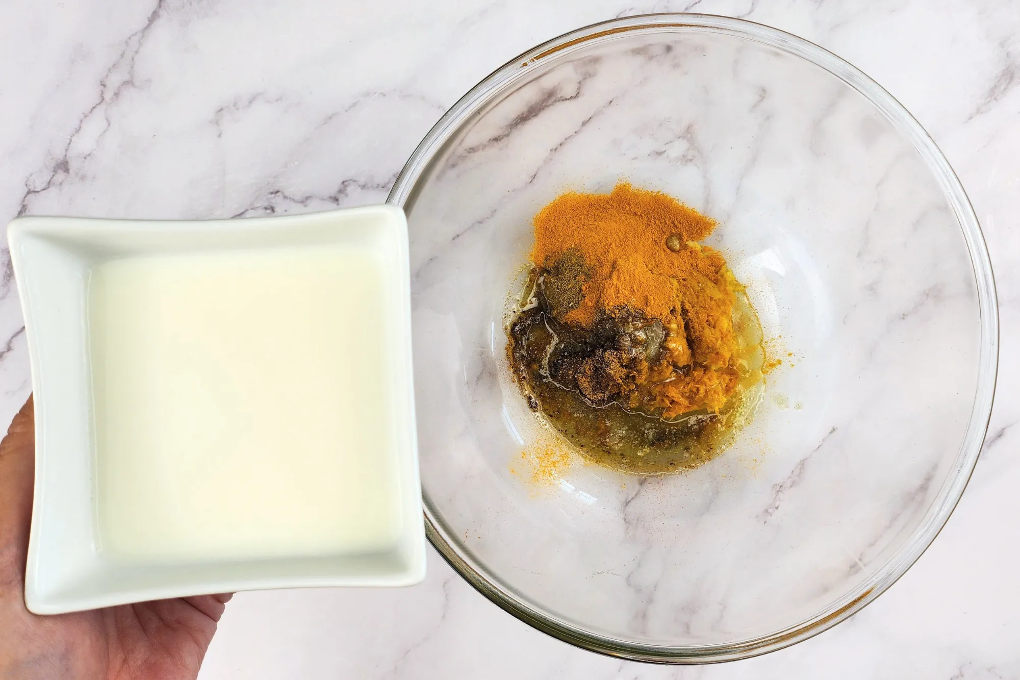 A hand is holding a bowl of milk above a mixing bowl with spices.