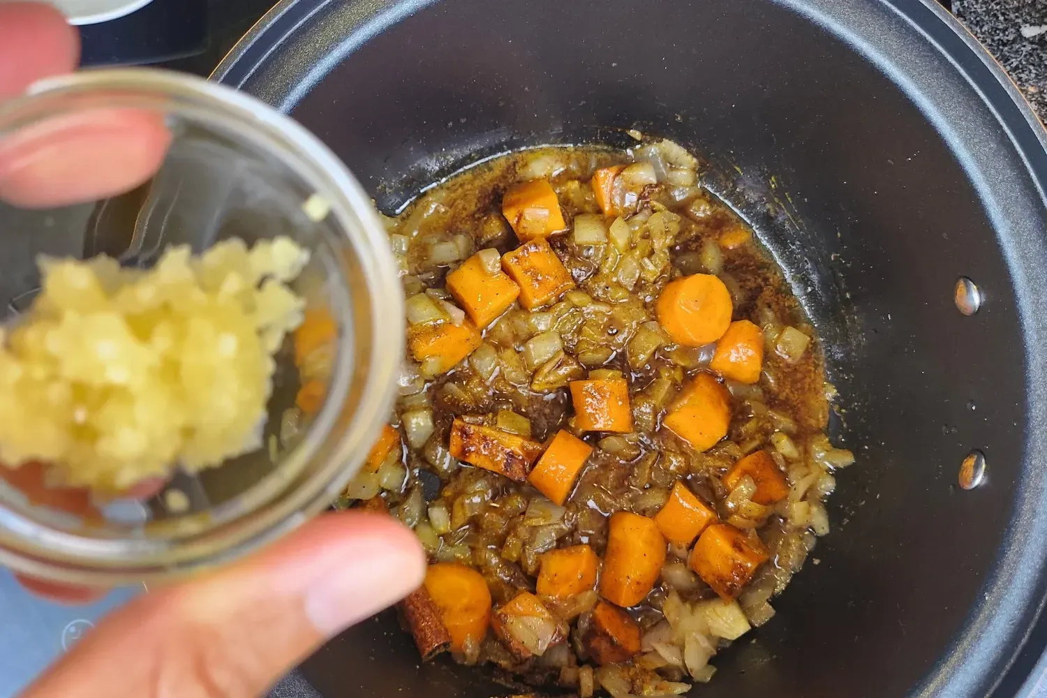 A hand is holding a bowl of minced garlic above a pot with cooked carrots and onions..
