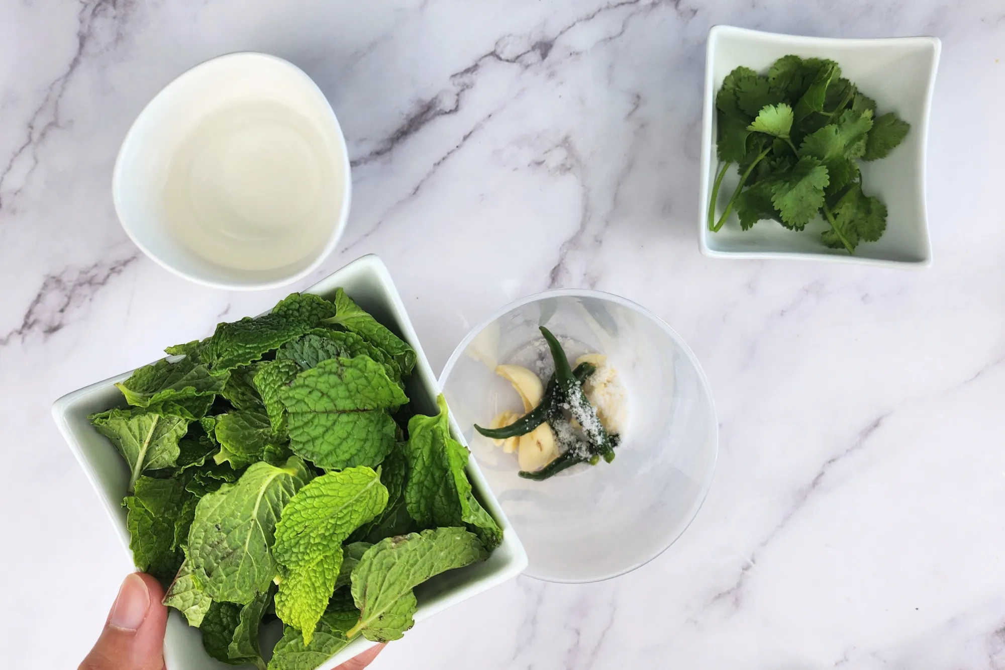A hand is holding a bowl of mint is above a mixing jar surrounded with cilantro, vinegar and honey.
