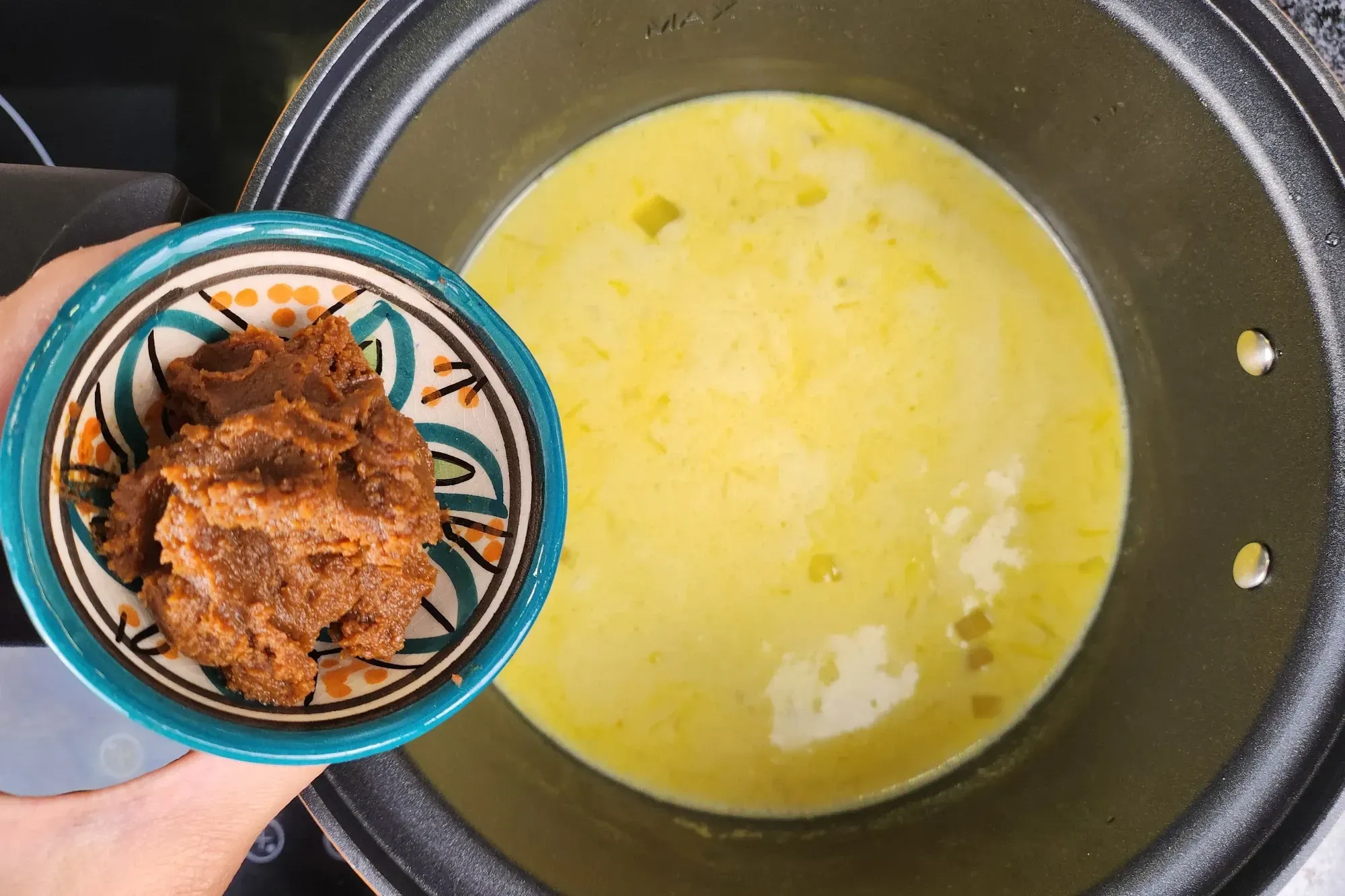 A hand is holding a bowl of miso paste above a pot