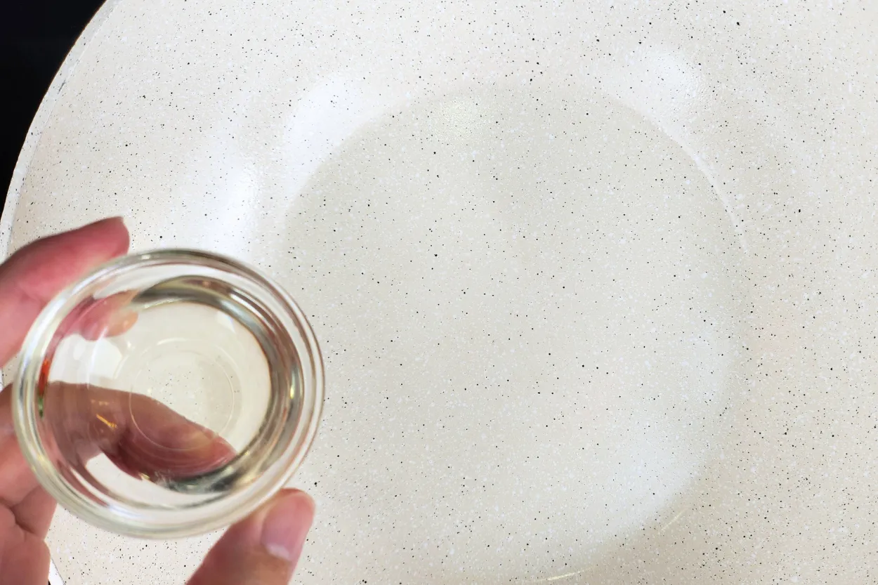 A hand is holding a bowl of oil above an empty wok.