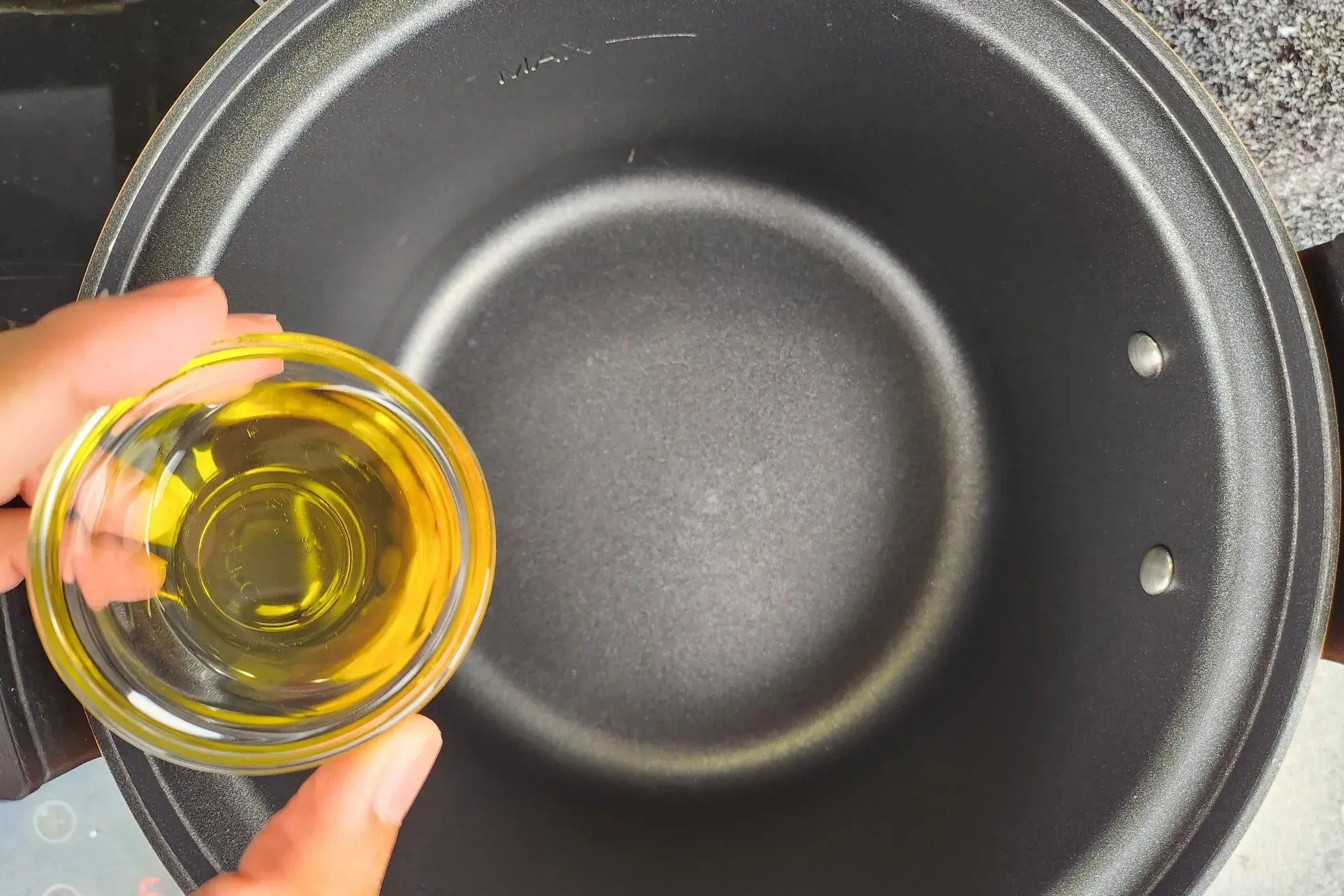 A hand is holding a bowl of olive oil above a black pot