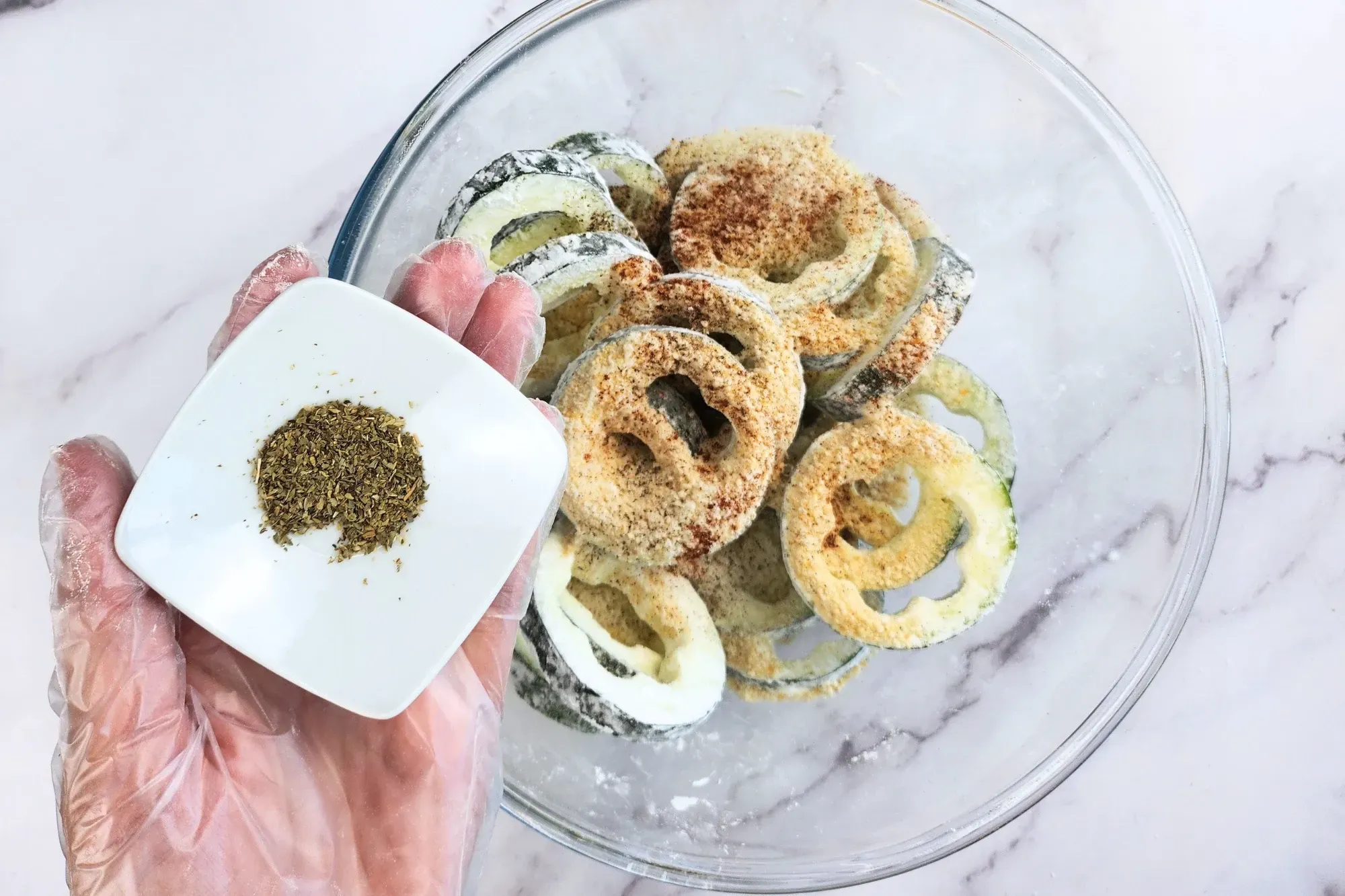 A hand is holding a bowl of origano above a bowl of zucchini rings