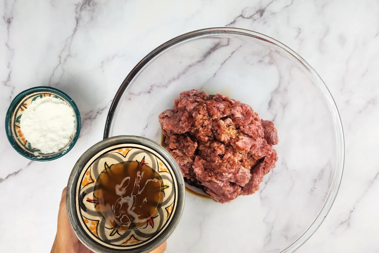 A hand is holding a bowl of oyster sauce above a mixing bowl with ground beef.