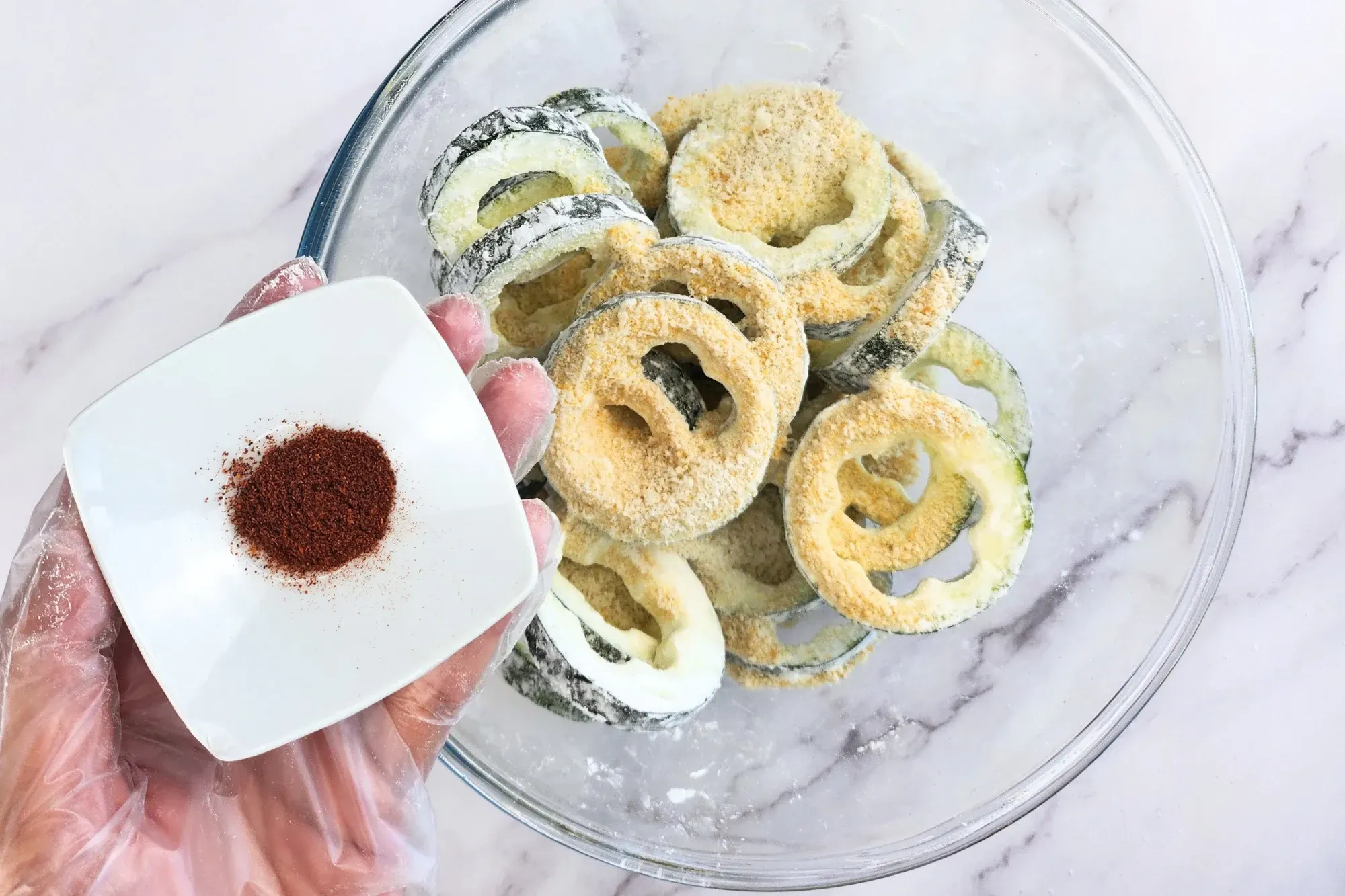A hand is holding a bowl of paprika above a bowl of zucchini rings