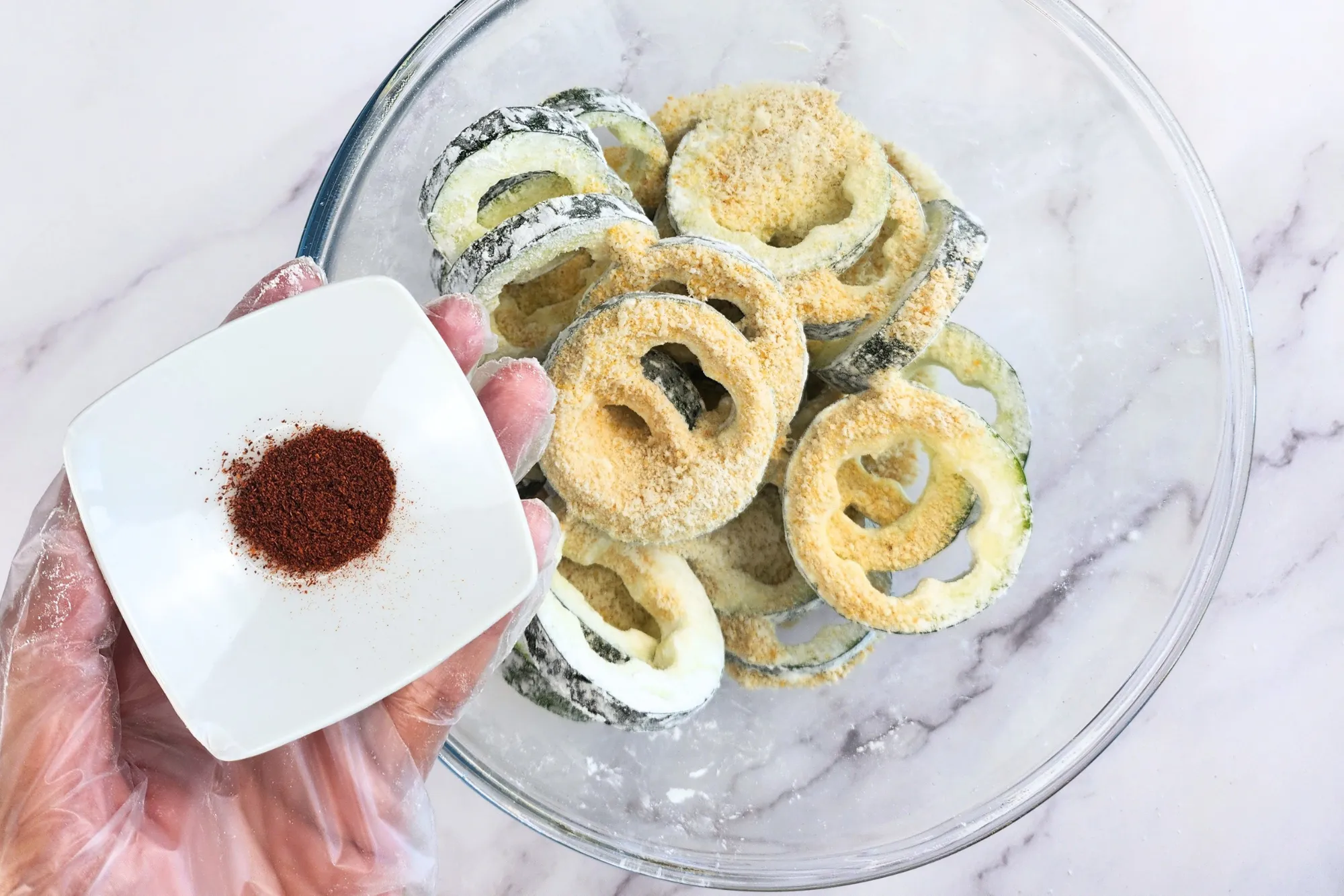 A hand is holding a bowl of paprika above a bowl of zucchini rings