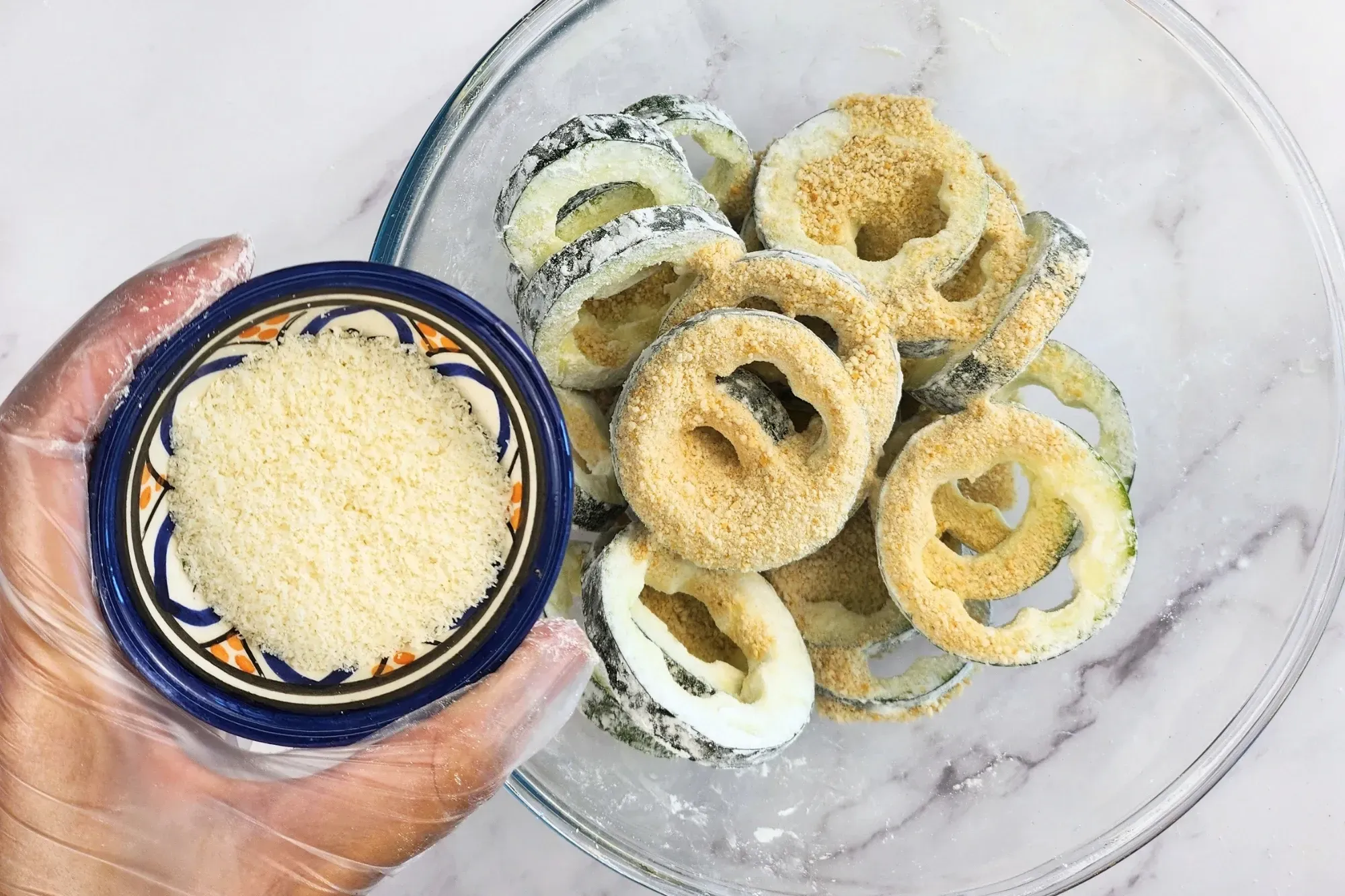 A hand is holding a bowl of parmesan above a bowl of zucchini rings.