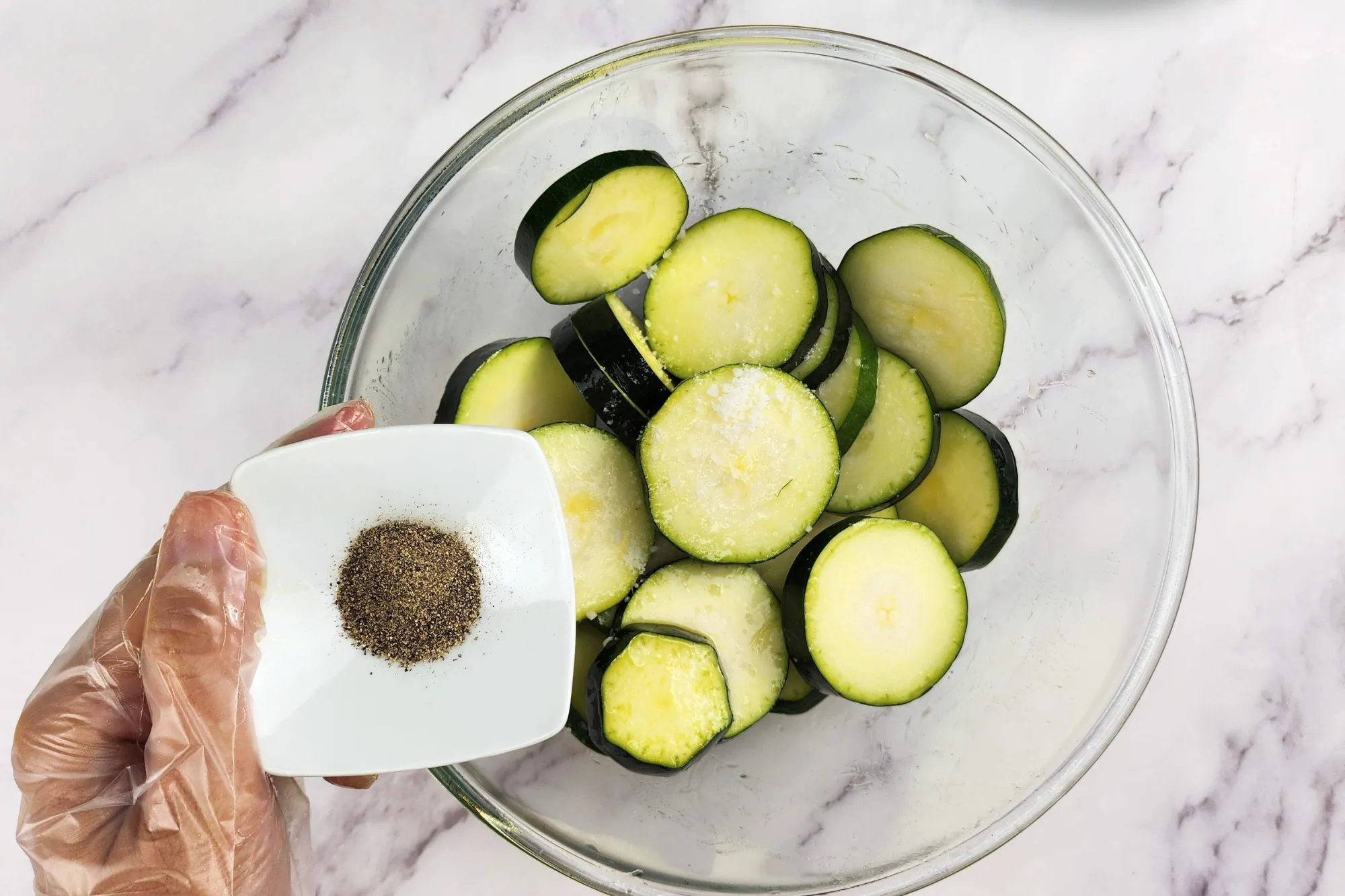 A hand is holding a bowl of pepper above a mixing bowl with zucchinis. 