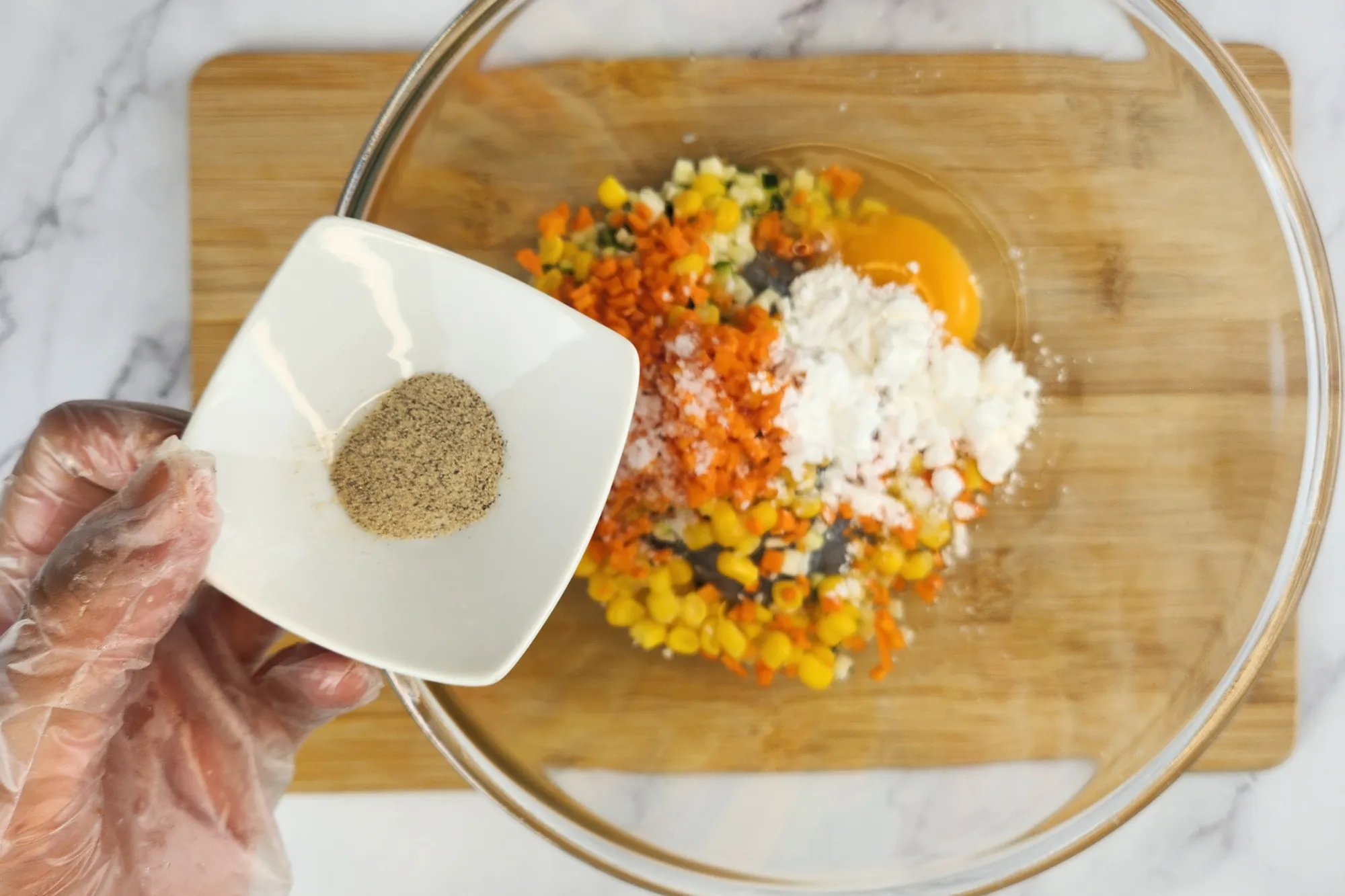 A hand is holding a bowl of pepper above a mixing bowl.