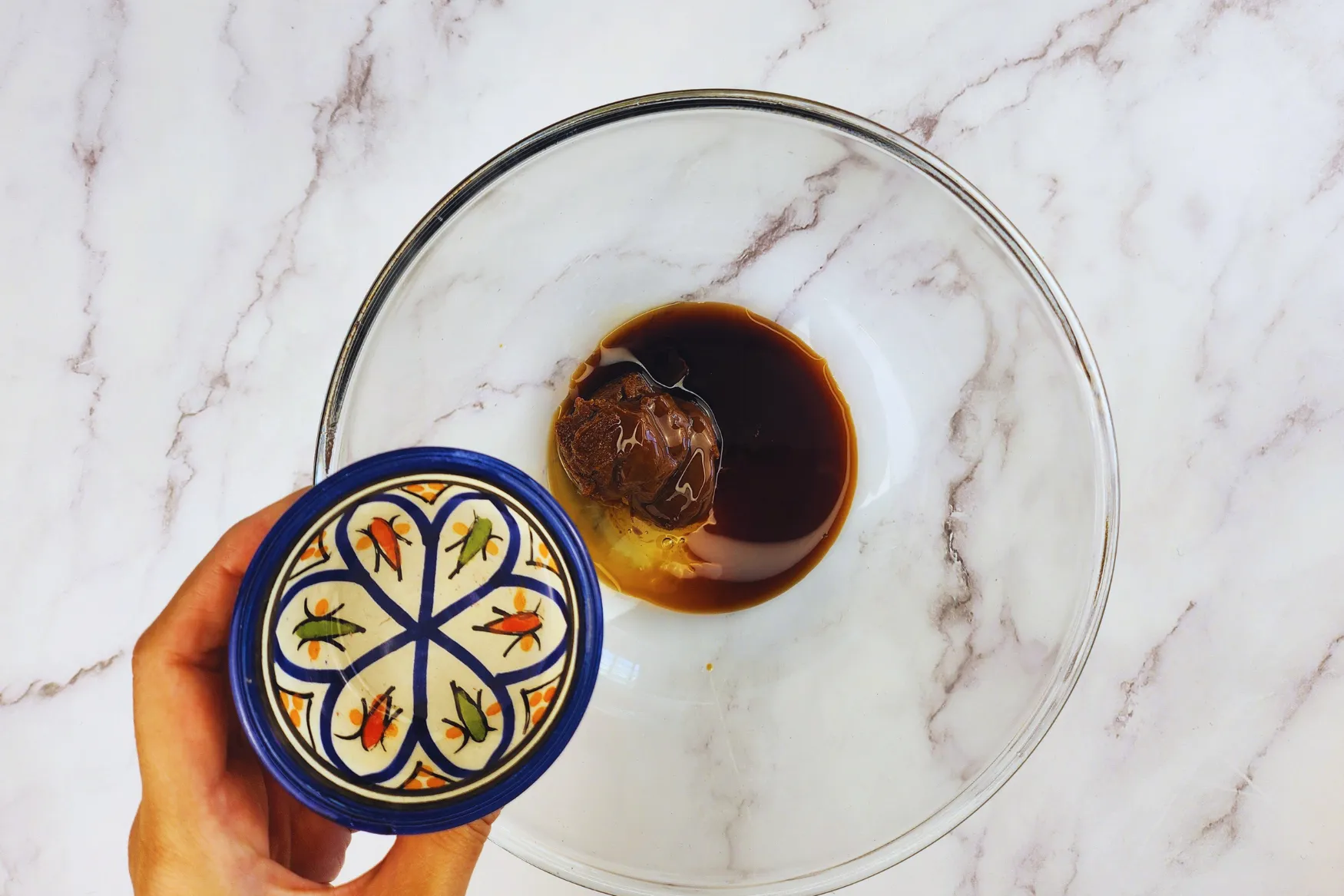 A hand is holding a bowl of rice vinegar above a mixing bowl.