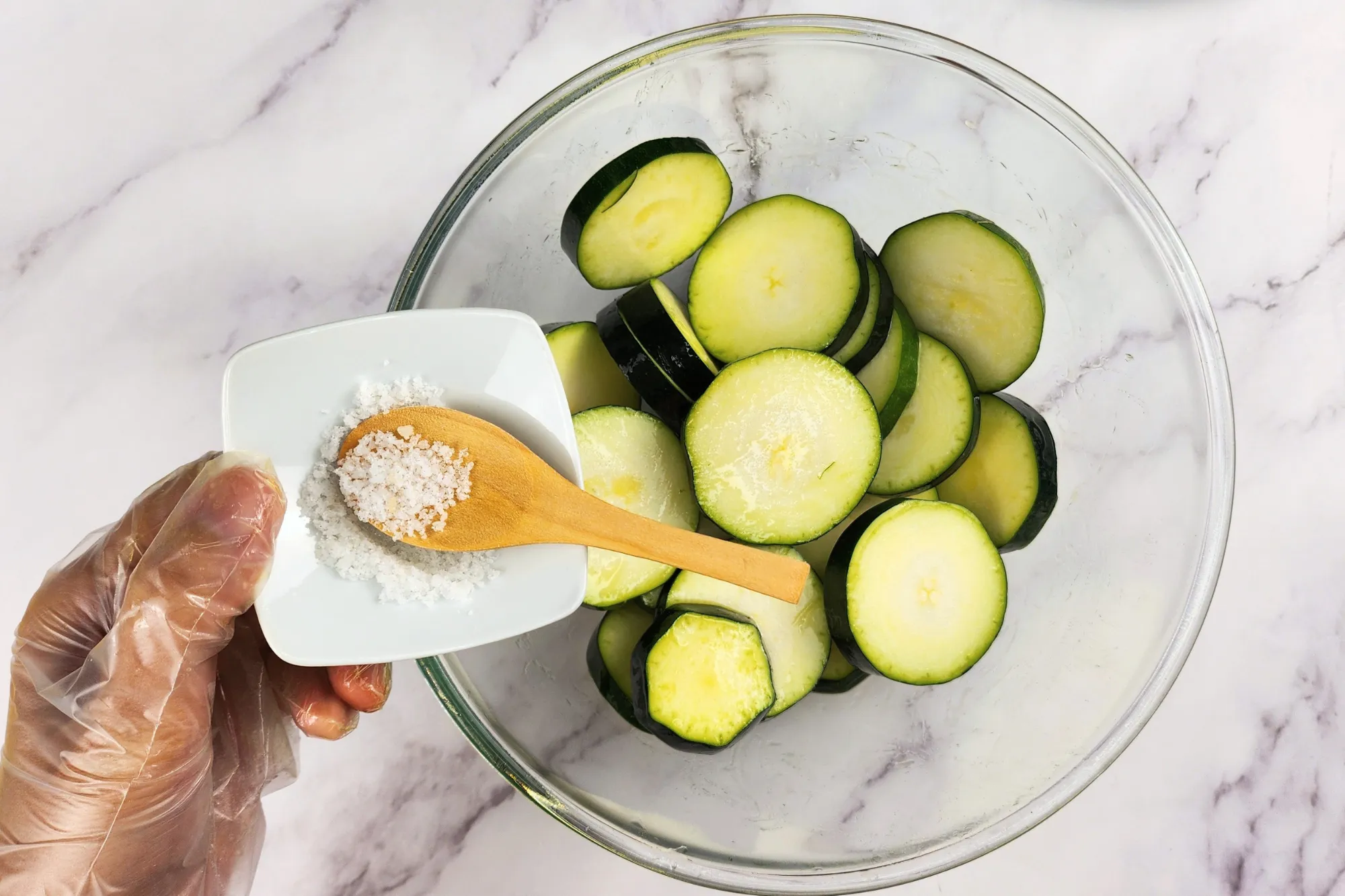 A hand is holding a bowl of salt above a mixing bowl with zucchinis. 