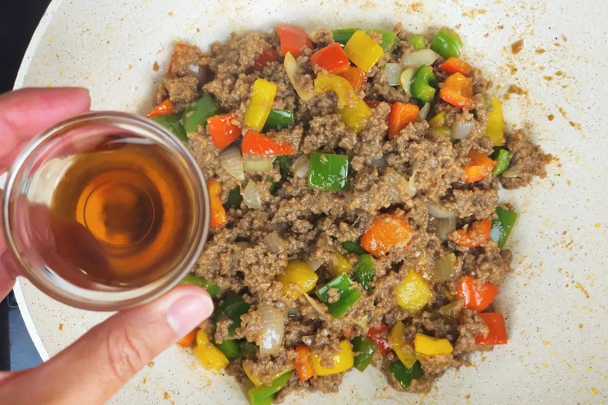 A hand is holding a bowl of sesame oil above cooked food in a wok.