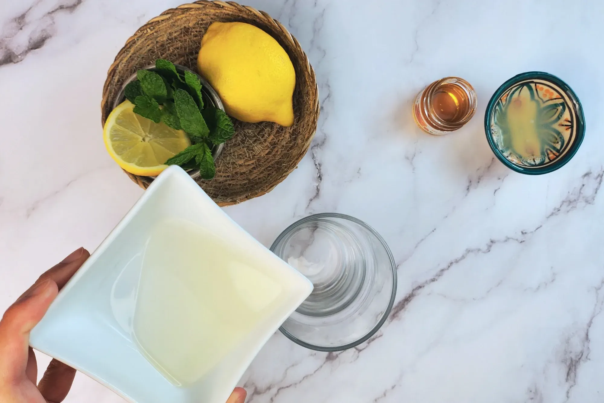 A hand is holding a bowl of syrup above a glass next to lemon, mint, honey and lemon juice.