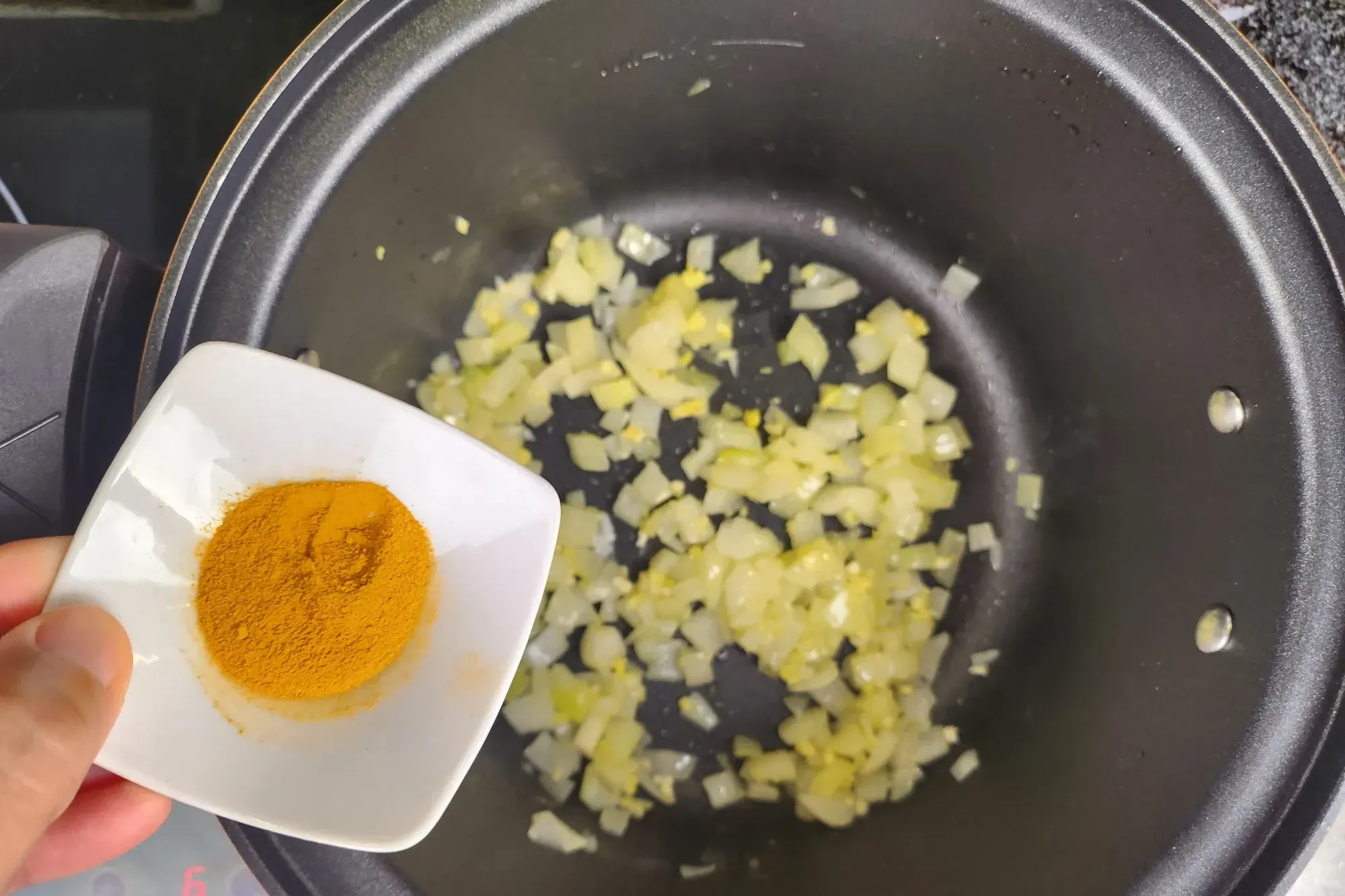 A hand is holding a bowl of turmeric powder garlic above a black pot with cooked onion
